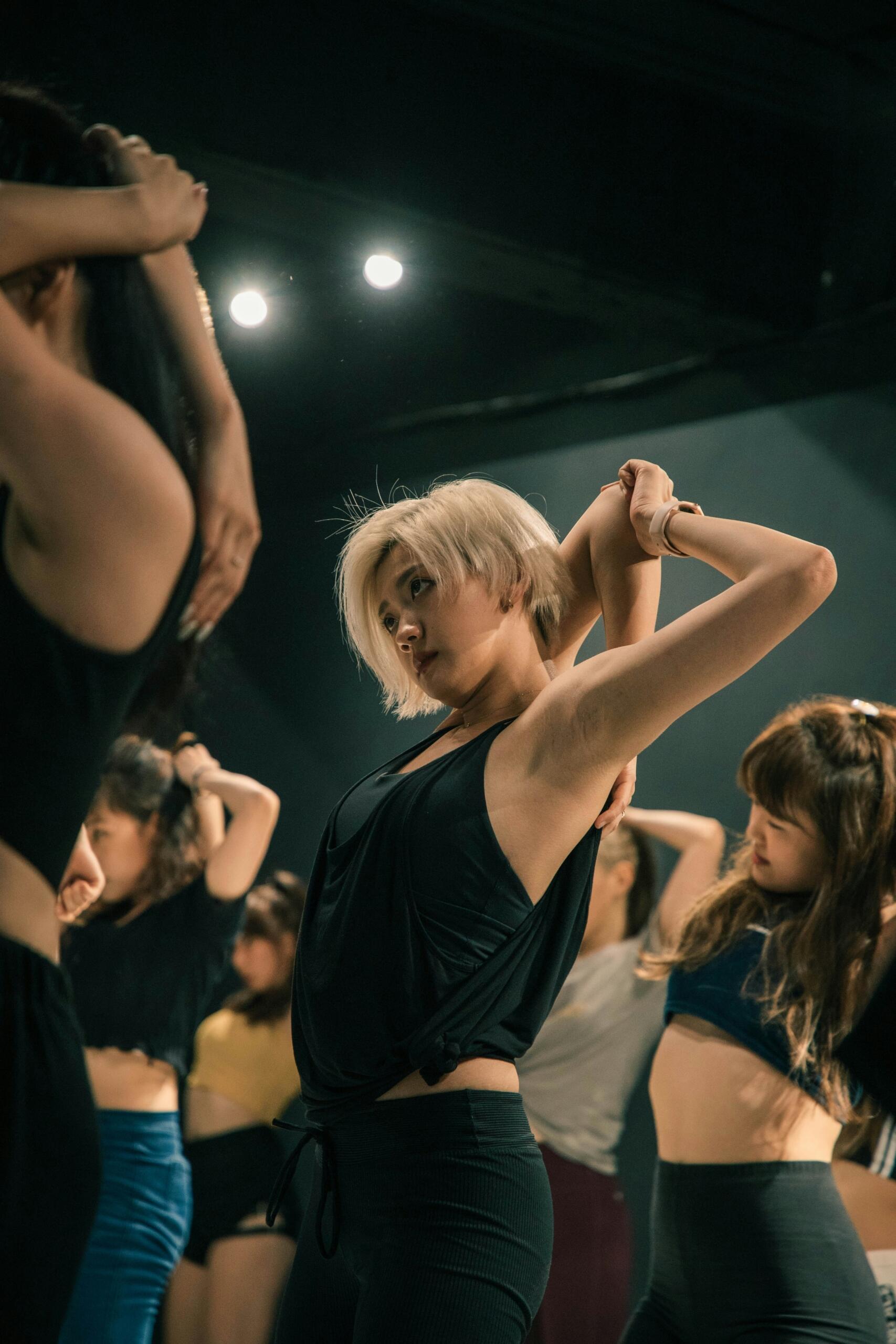A group of women in athletic wear stretching their arms behind their heads in a dimly lit indoor exercise or dance studio.