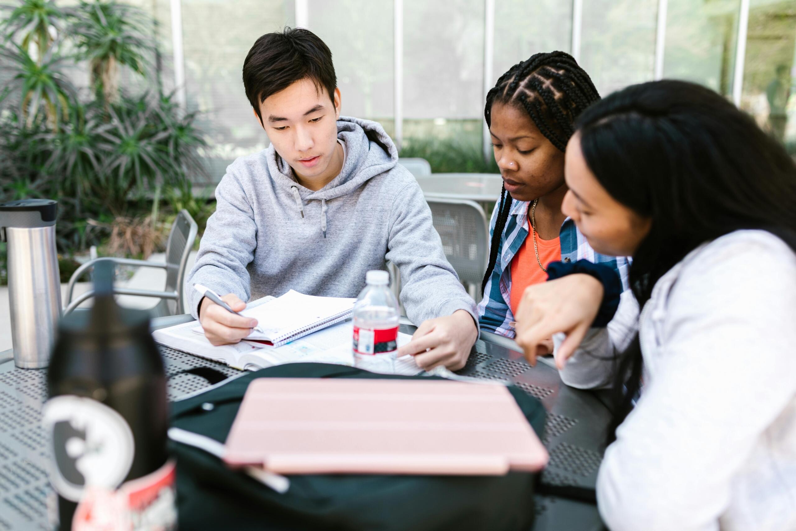 small group of students sits around a table studying together while a tutor provides guidance and support. Open textbooks, notebooks, and learning materials are spread across the table as the students listen attentively and collaborate on their work in a positive academic environment.