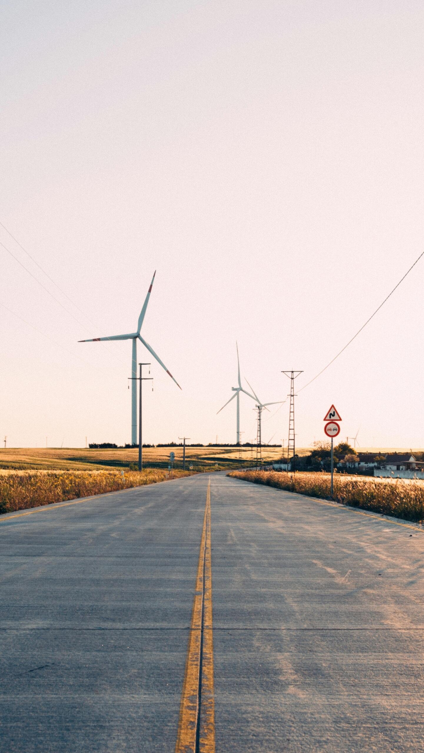 Several modern wind turbines stand across an open landscape, their large white blades turning in the wind. The turbines rise above fields under a clear sky, generating renewable energy through wind power.