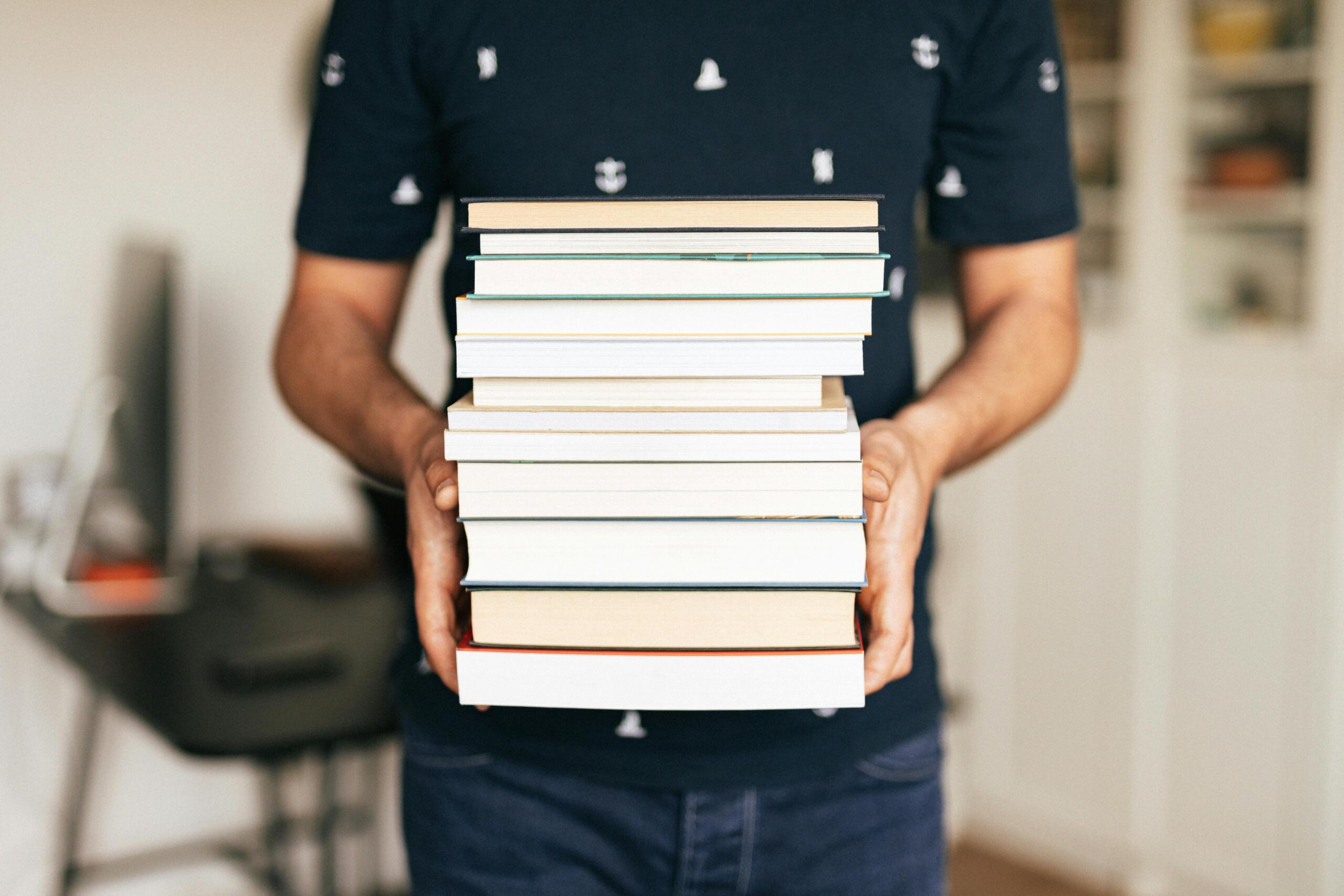 A student holds a stack of textbooks and notebooks in front of their body. Their face is not visible, and the books are the main focus of the image, suggesting studying, learning, or academic preparation.