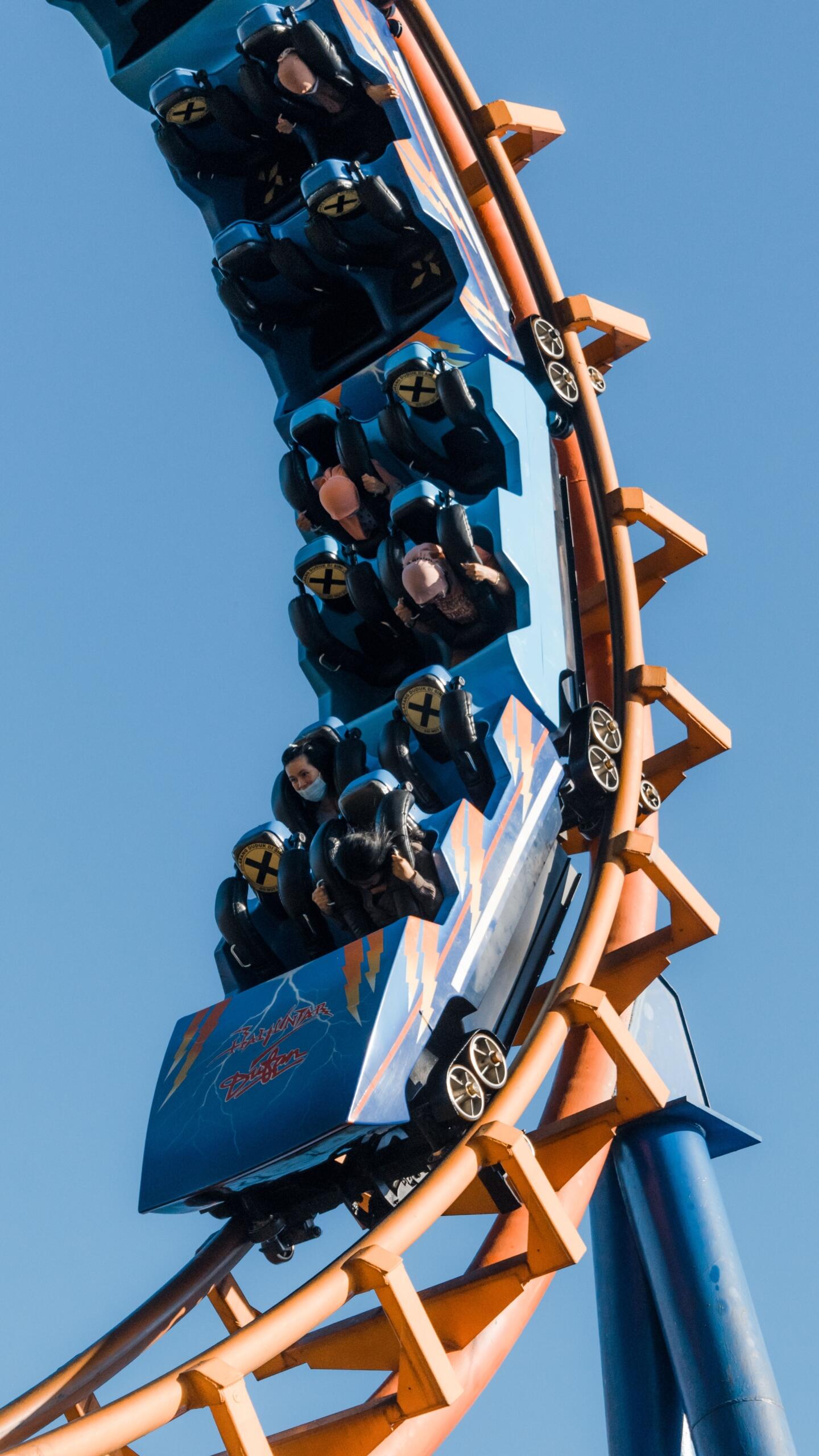 A roller coaster train speeds along a steep track with riders raising their hands as it climbs and descends hills. The image illustrates key physics concepts such as gravity, potential energy, kinetic energy, acceleration, and motion as the coaster moves through the course.