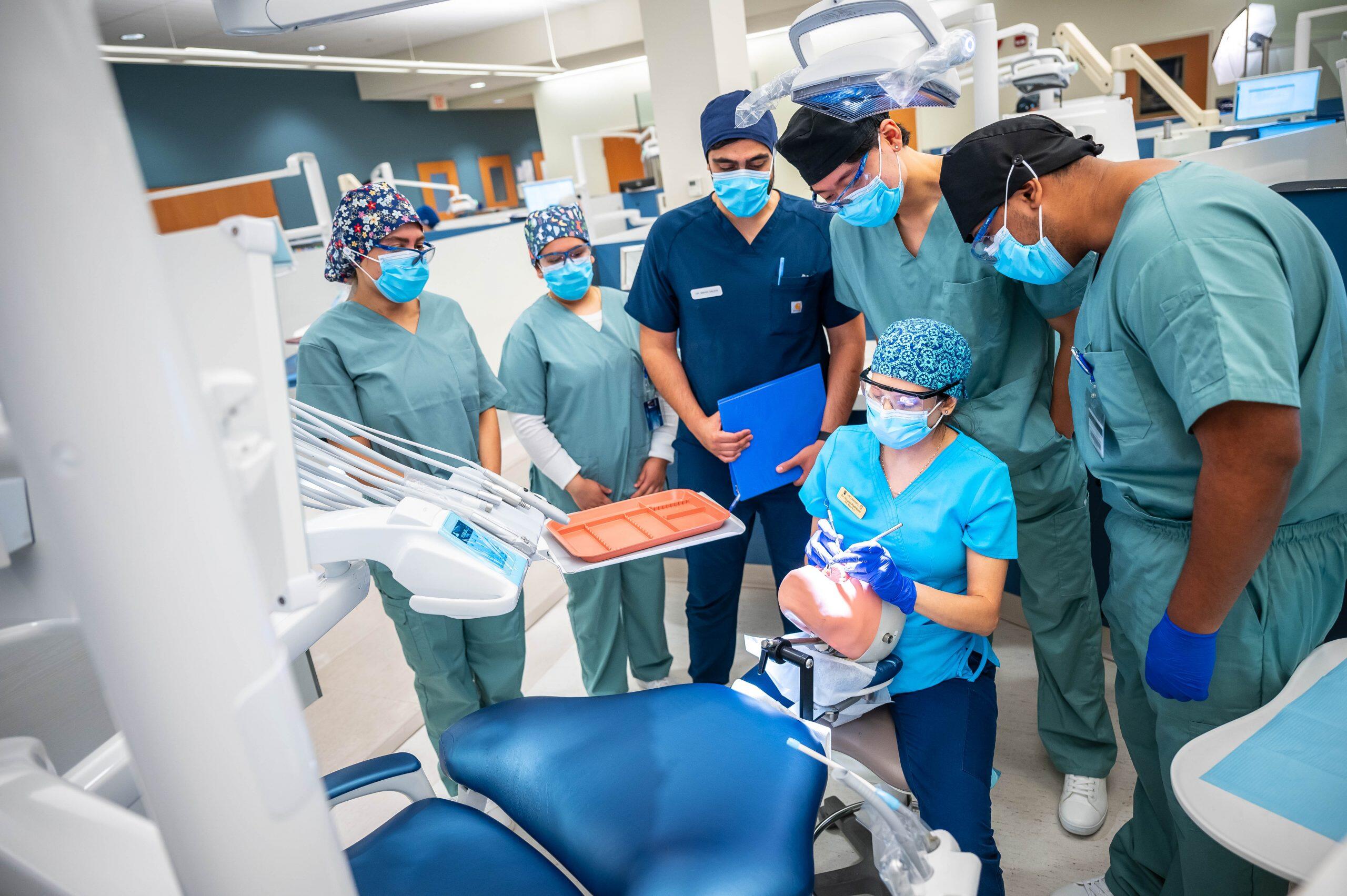 Dental students in scrubs observe a hands-on training session using a dental mannequin in a modern clinic setting.