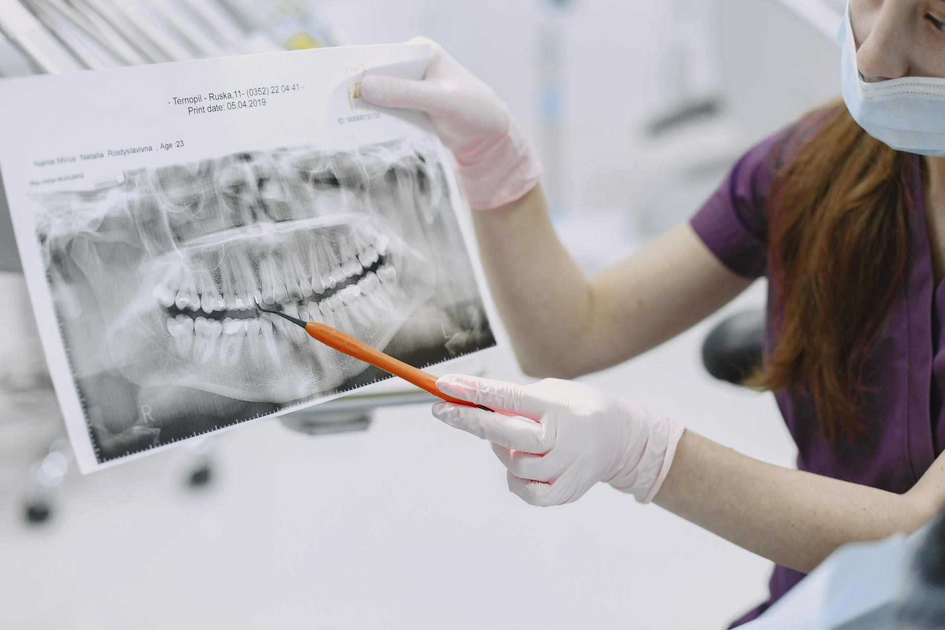 Dentist wearing gloves and mask points at dental X-ray with a pen, explaining teeth condition to a patient.