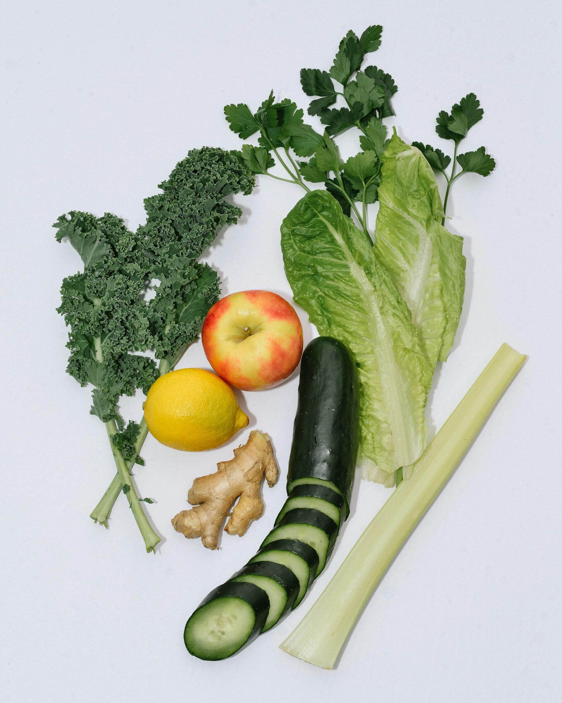 Fresh ingredients on white background including kale, parsley, apple, lemon, ginger, romaine lettuce, sliced cucumber, and celery stalk.