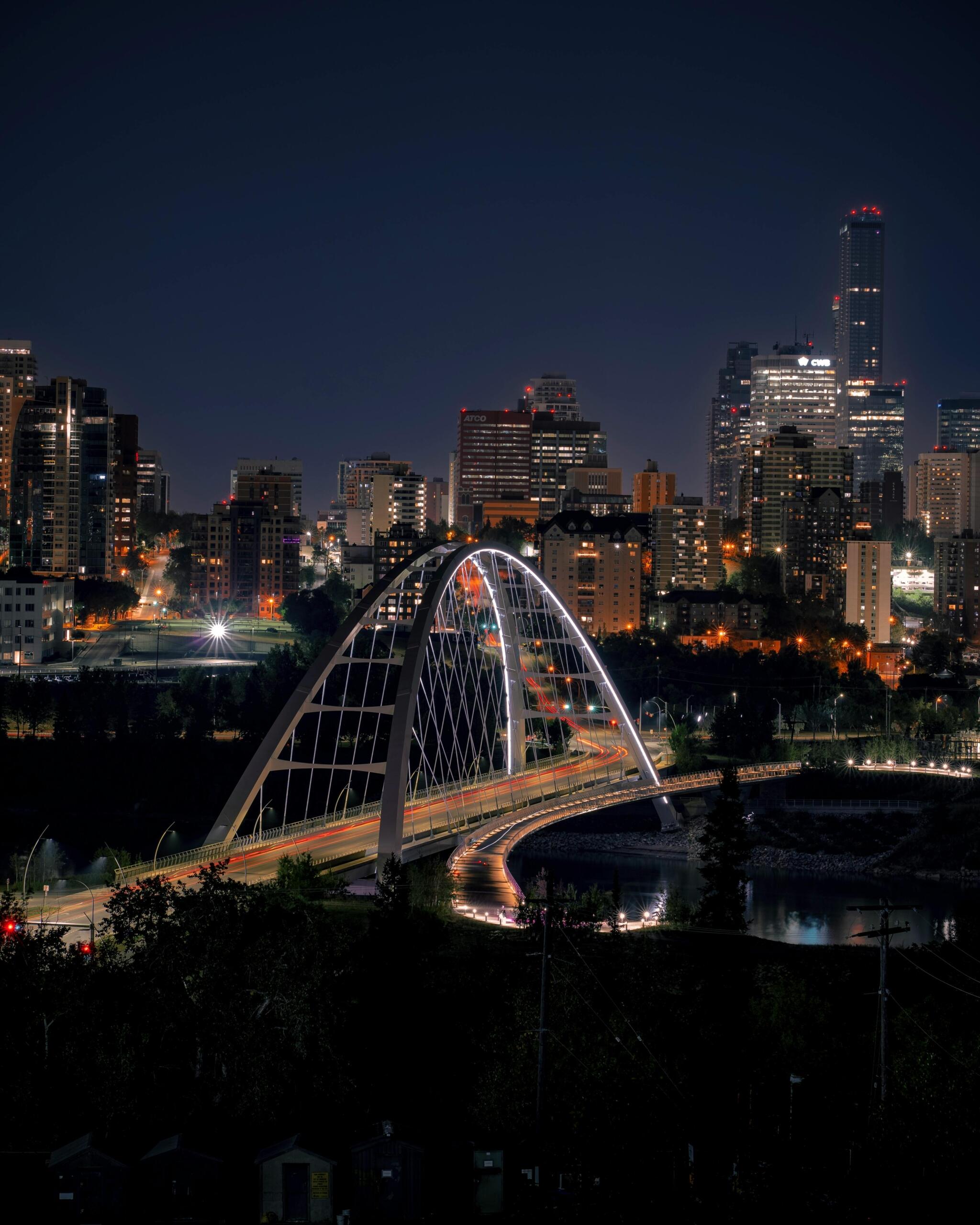 Skyline of Edmonton at night with illuminated high-rise buildings, glowing streetlights, and reflections along the river valley under a dark sky.