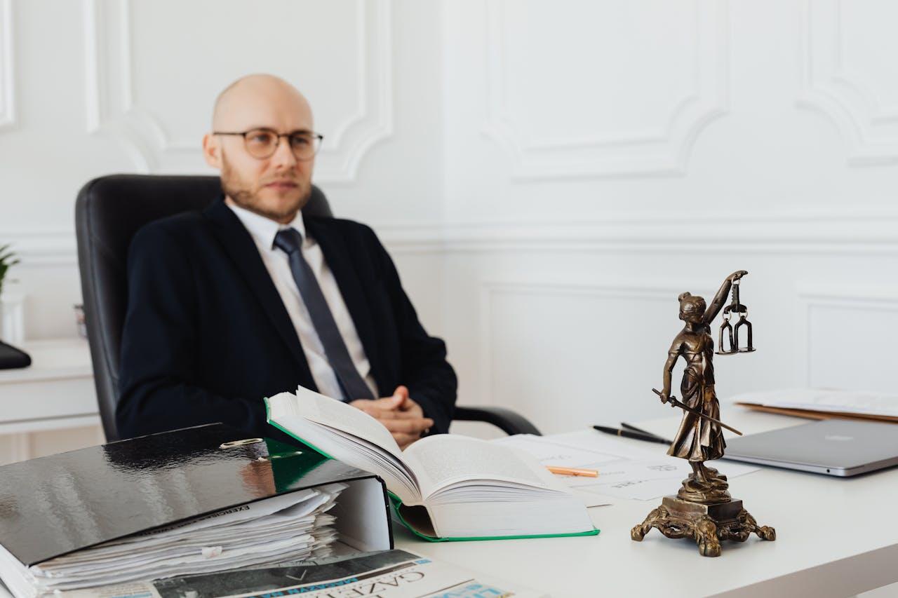 A man in a suit sits at a desk, with a statue of Lady Justice prominently displayed beside him.