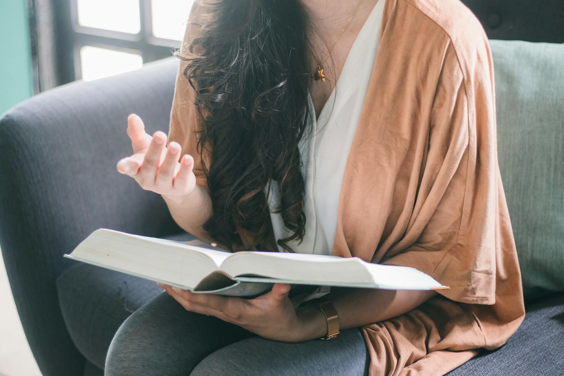 Person sitting on a couch, reading a large book, and gesturing with one hand, wearing a brown shawl and white shirt.