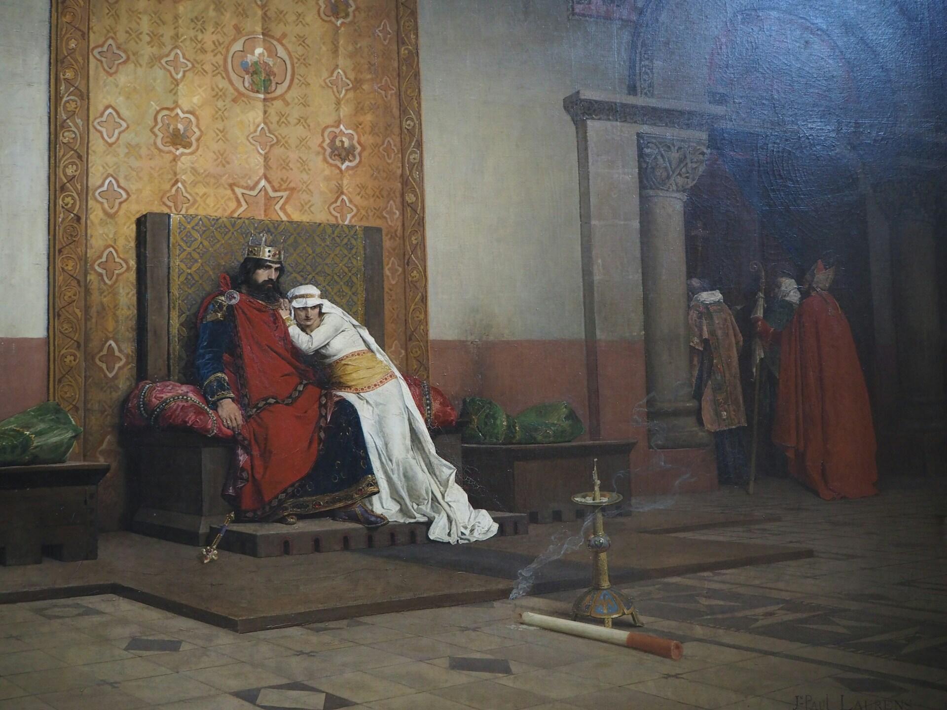 A medieval king seated on a throne, embraced by a woman in white, with attendants in red cloaks near stone columns in a grand hall.