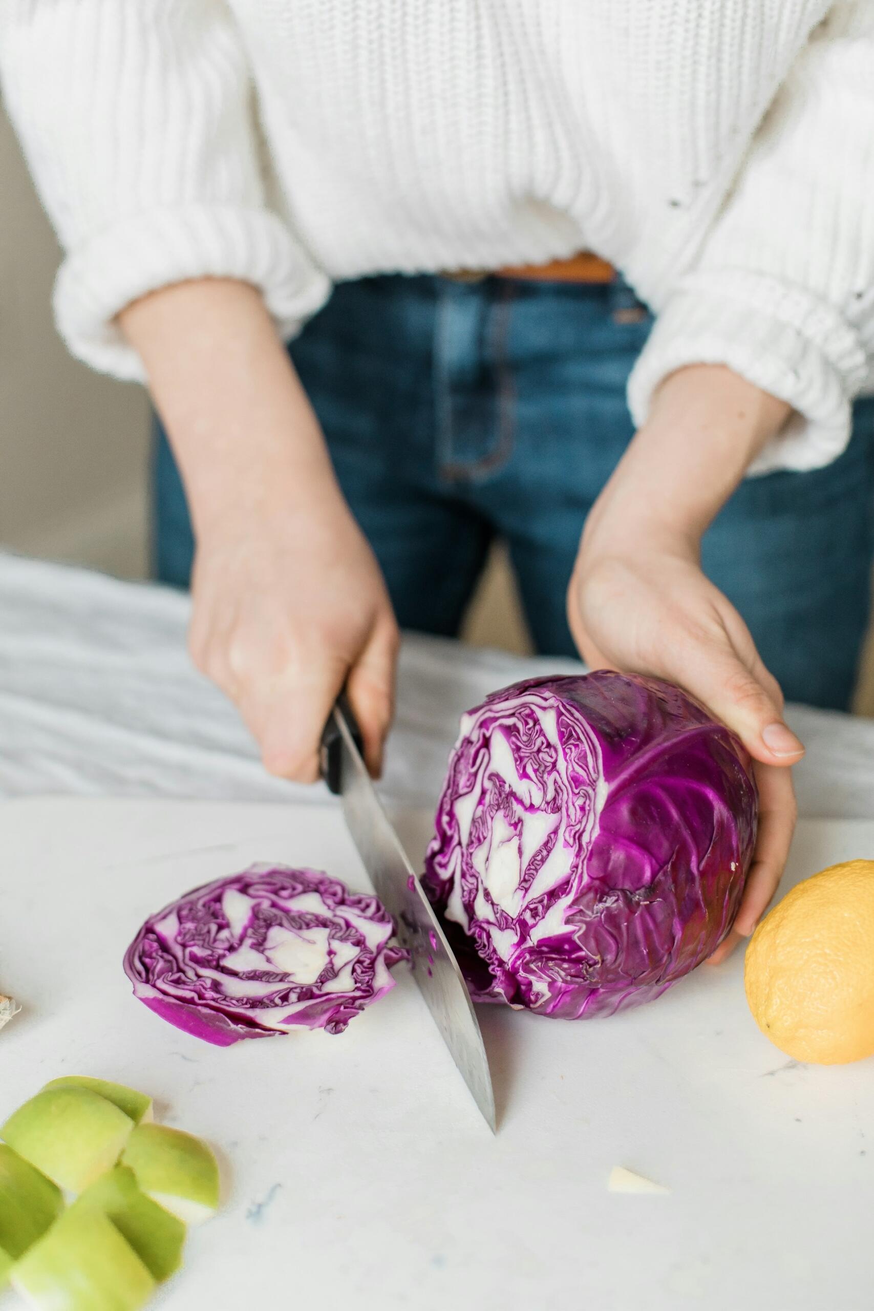 Person slicing a purple cabbage on a white surface, with cut apple pieces and a lemon nearby.