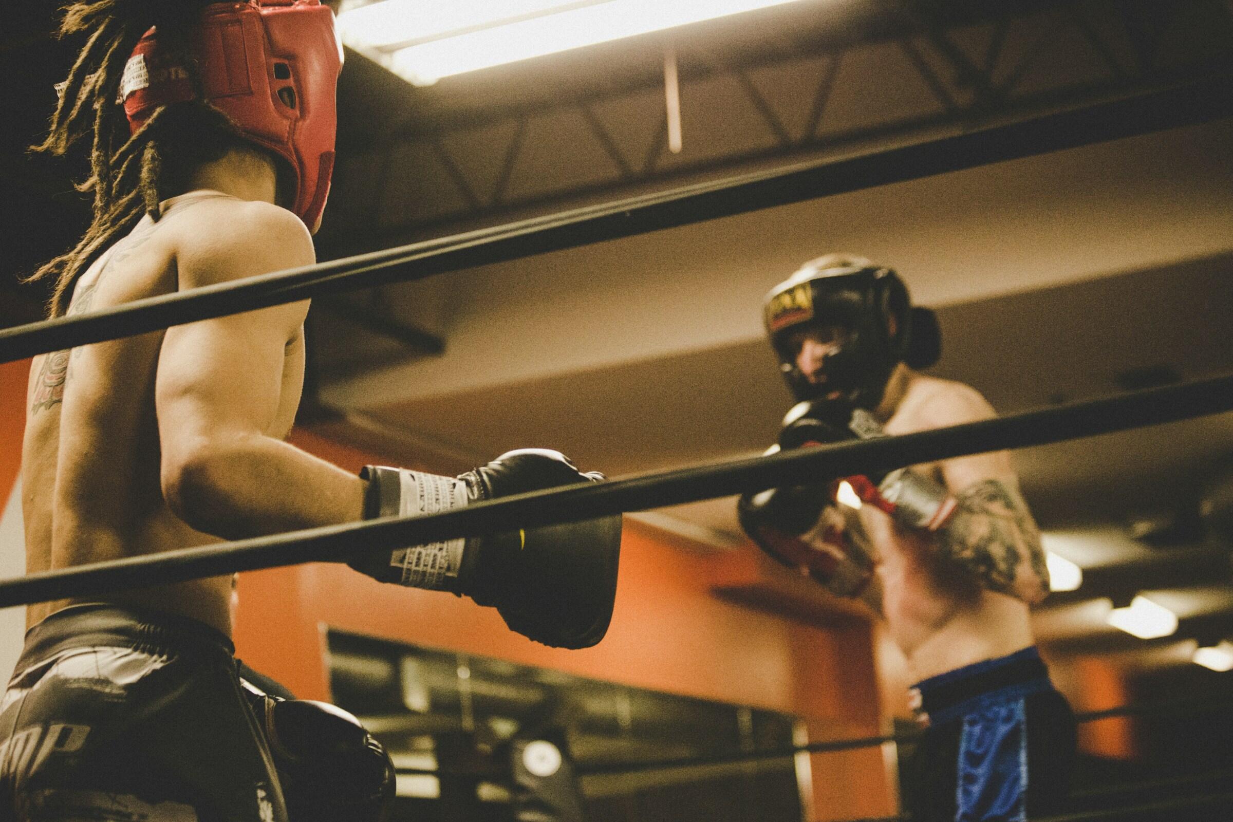 Two shirtless boxers wearing protective headgear and gloves face off inside a boxing ring in a gym.