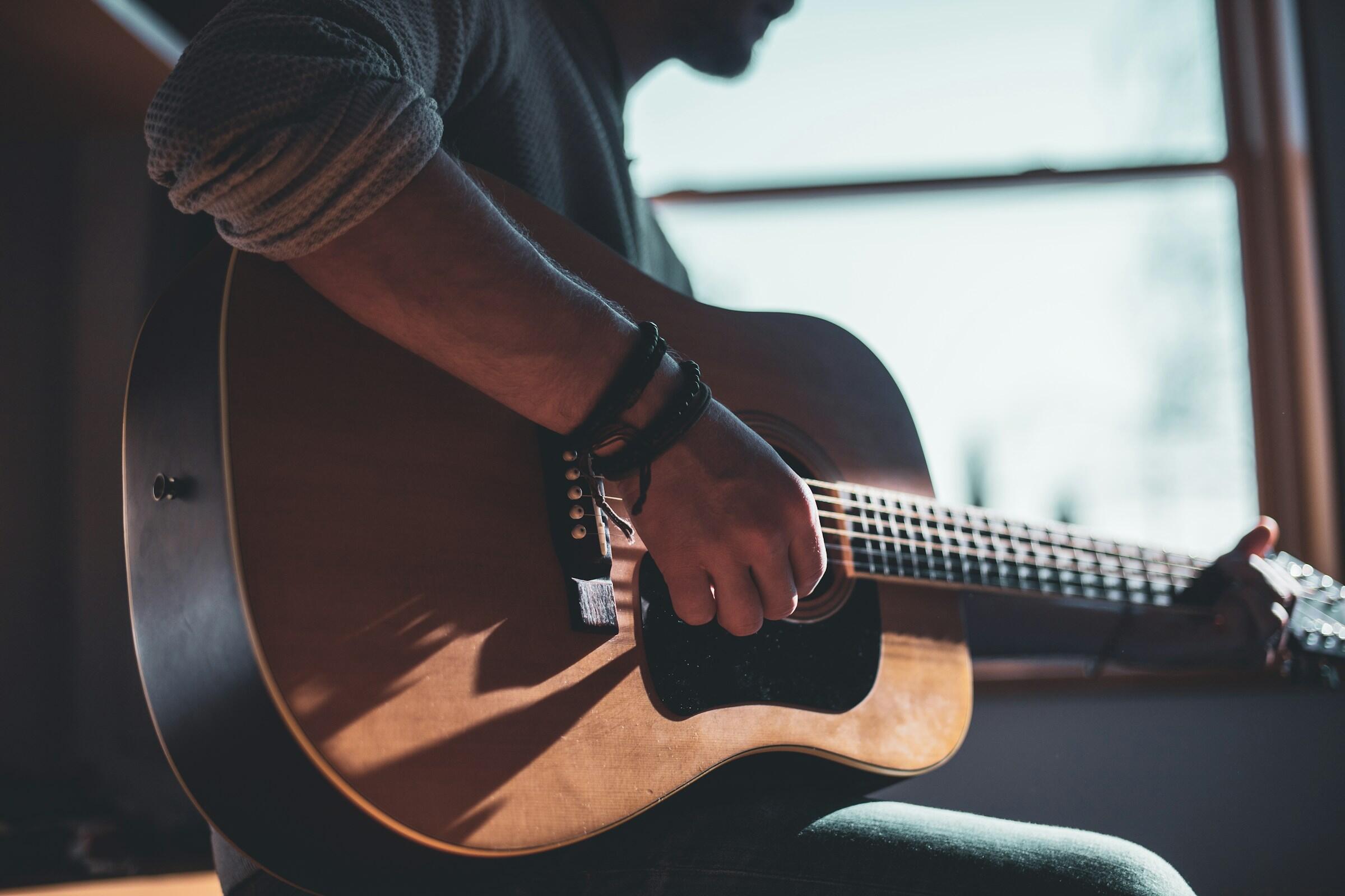 Person playing an acoustic guitar indoors near a sunlit window, focusing on the guitar and hands strumming the strings.
