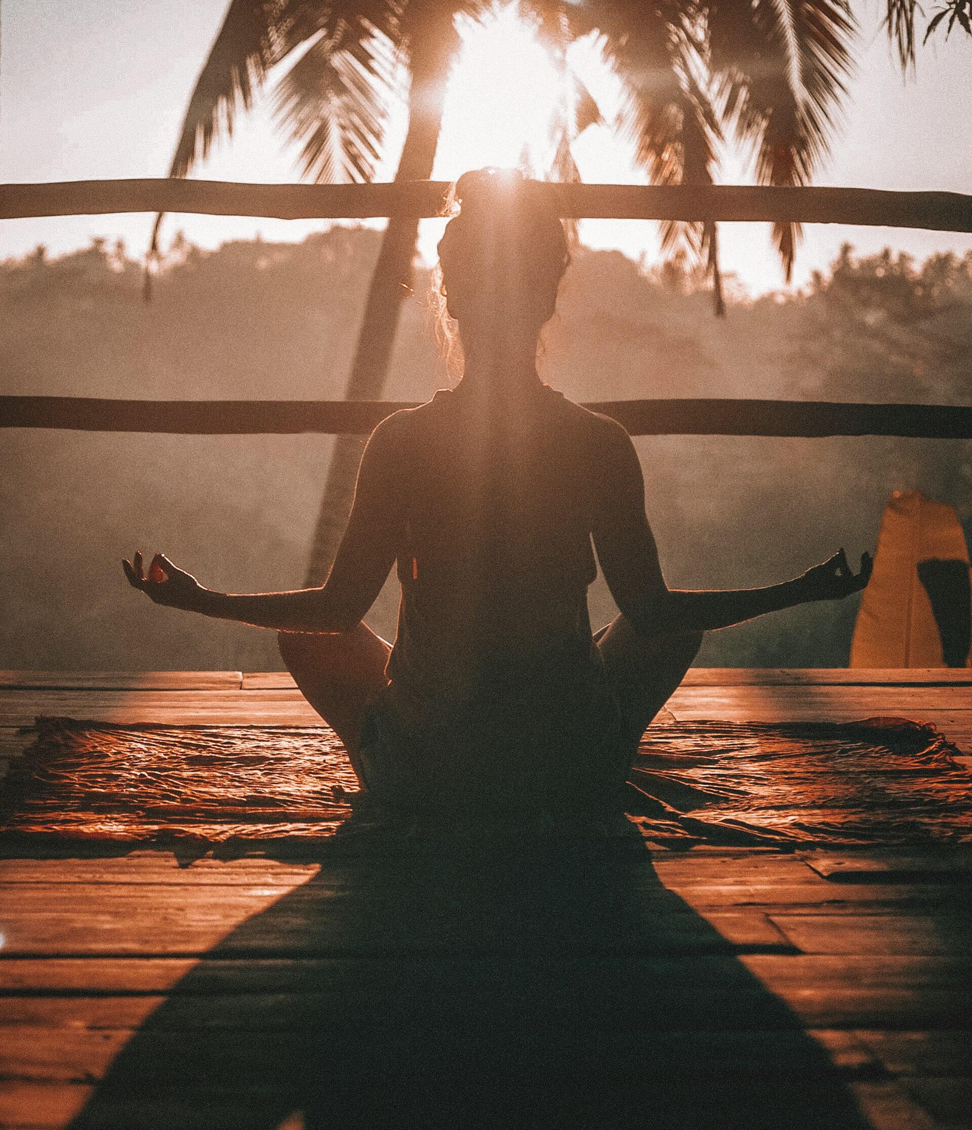 Silhouette of a person meditating in lotus pose on a wooden deck at sunrise, with palm trees and warm sunlight in the background.