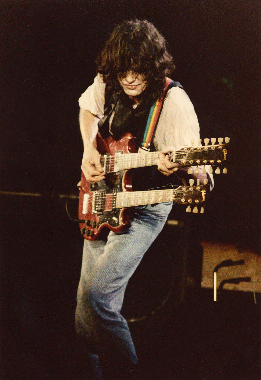 Musician playing a double-neck electric guitar on stage with a rainbow guitar strap, dressed in a white shirt and blue jeans.