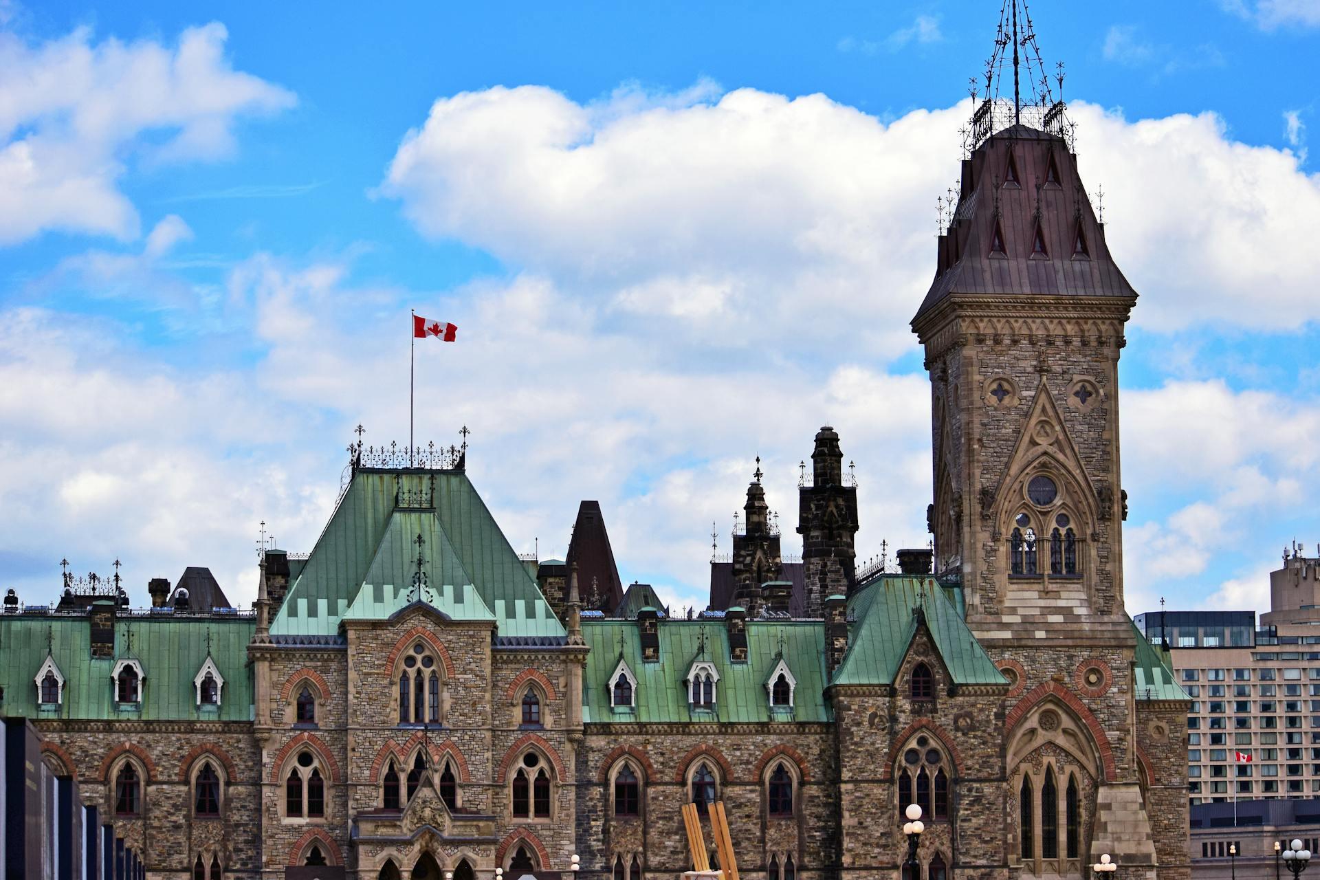 Historic stone building with green roofs and a tall tower under a partly cloudy sky, flying the Canadian flag.
