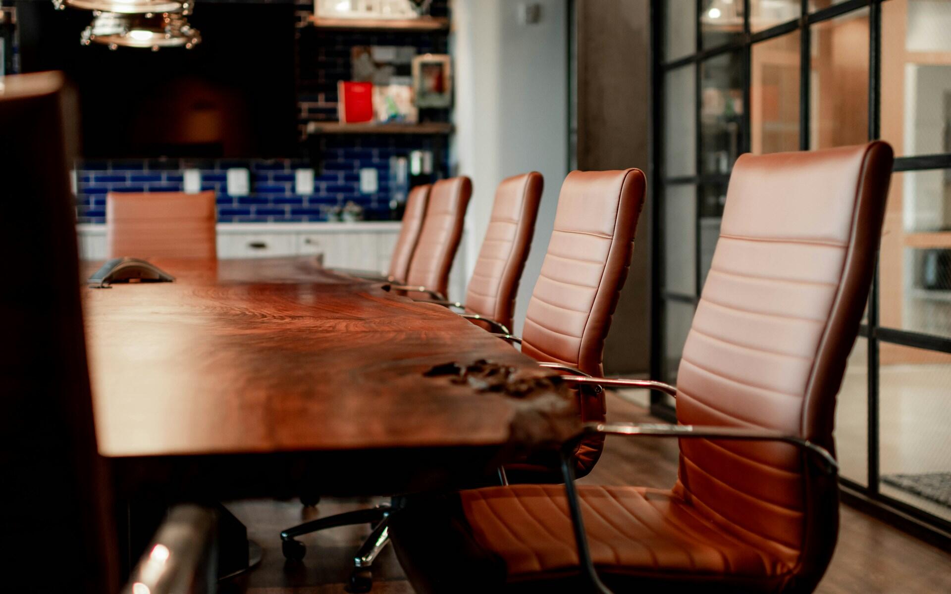 Modern conference room with a long wooden table and several brown leather office chairs arranged around it.