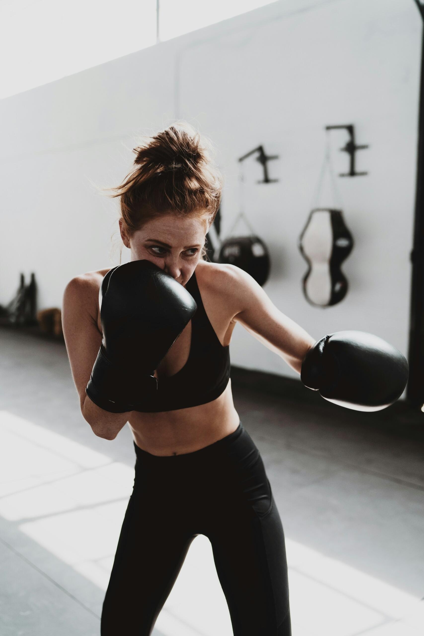Woman in black sportswear and boxing gloves practicing punches in a well-lit gym with punching bags on the wall.