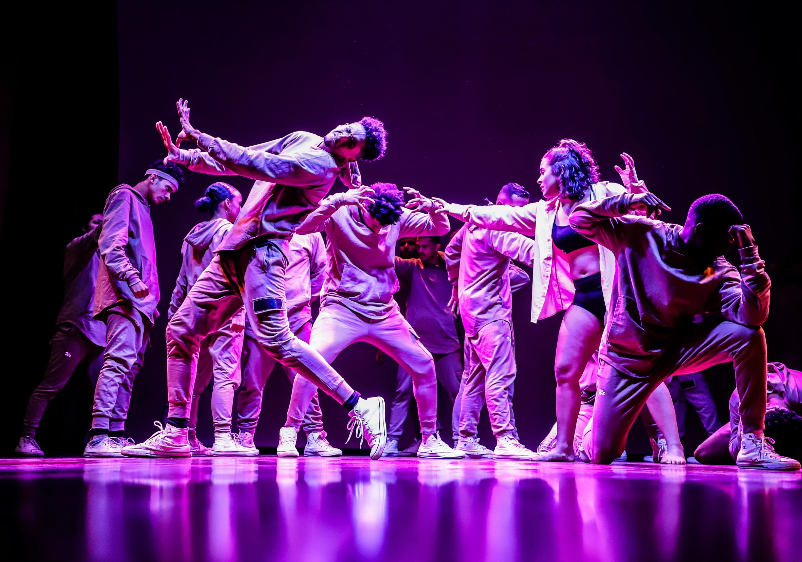 A group of dancers in casual attire striking dynamic poses on stage under vibrant purple lighting.