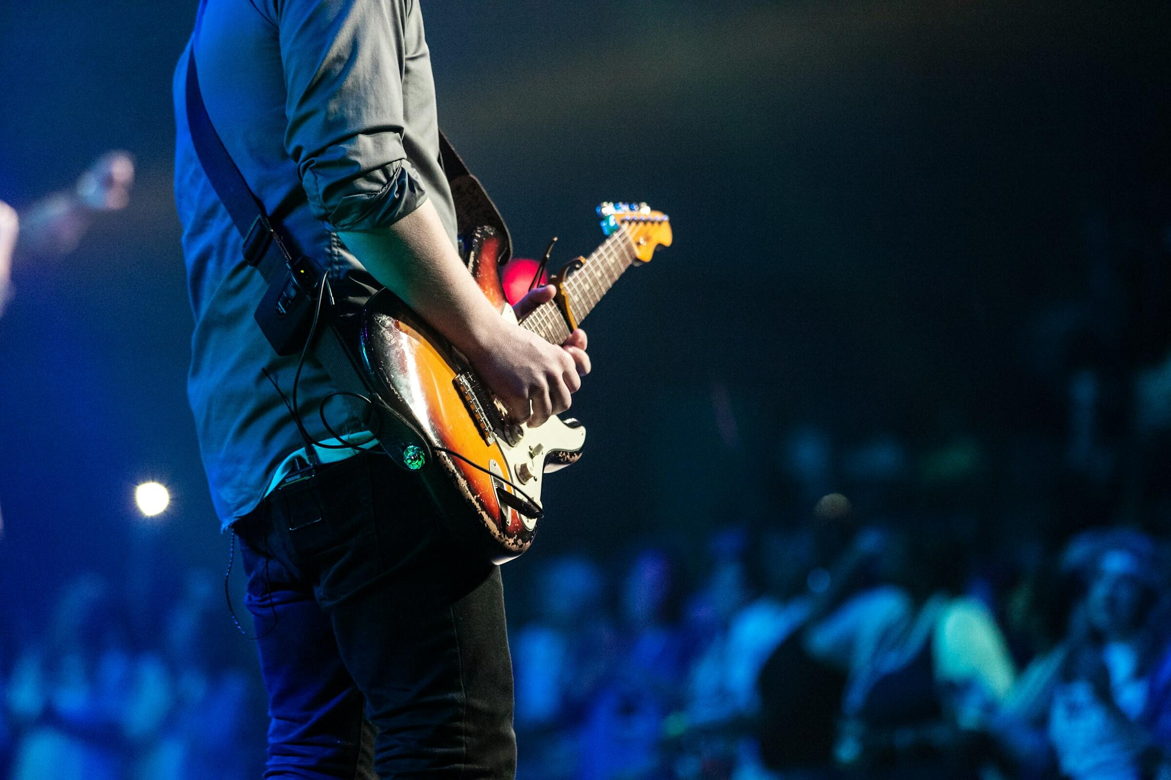 Musician playing electric guitar on stage with an audience blurred in blue lighting in the background.