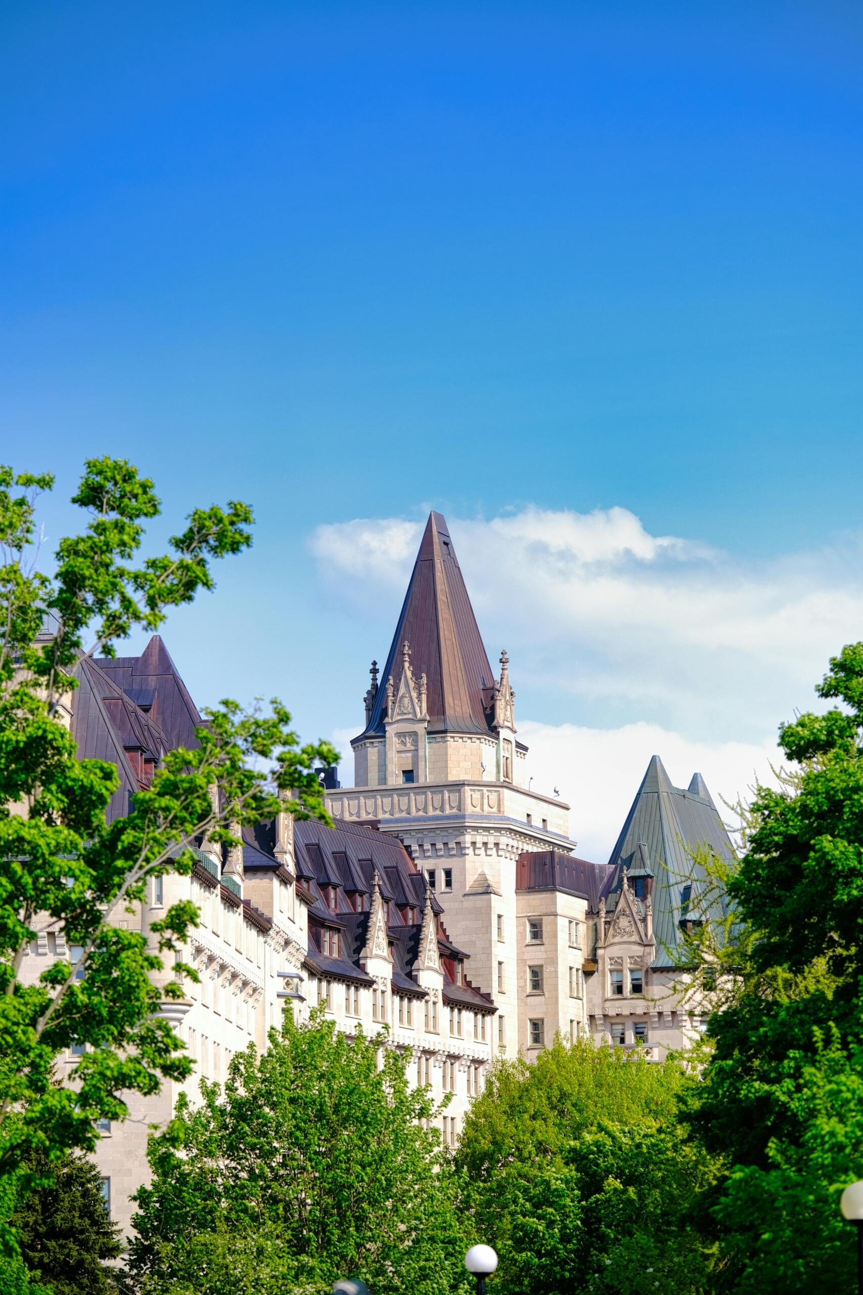 A grand château-style hotel in Ottawa with a steep copper roof, ornate stone façade, and turreted towers, overlooking the Ottawa River and nearby historic landmarks under a clear sky.