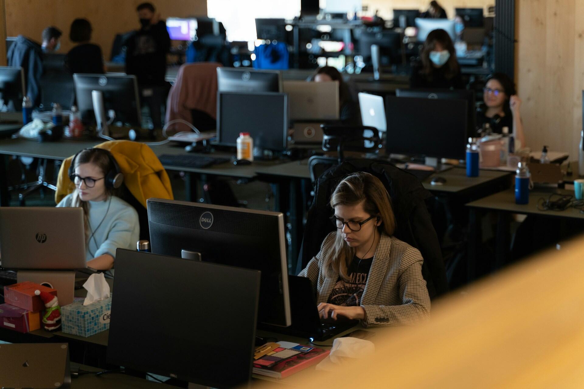 Open office with people working at computers, some wearing headphones and one person wearing a face mask.