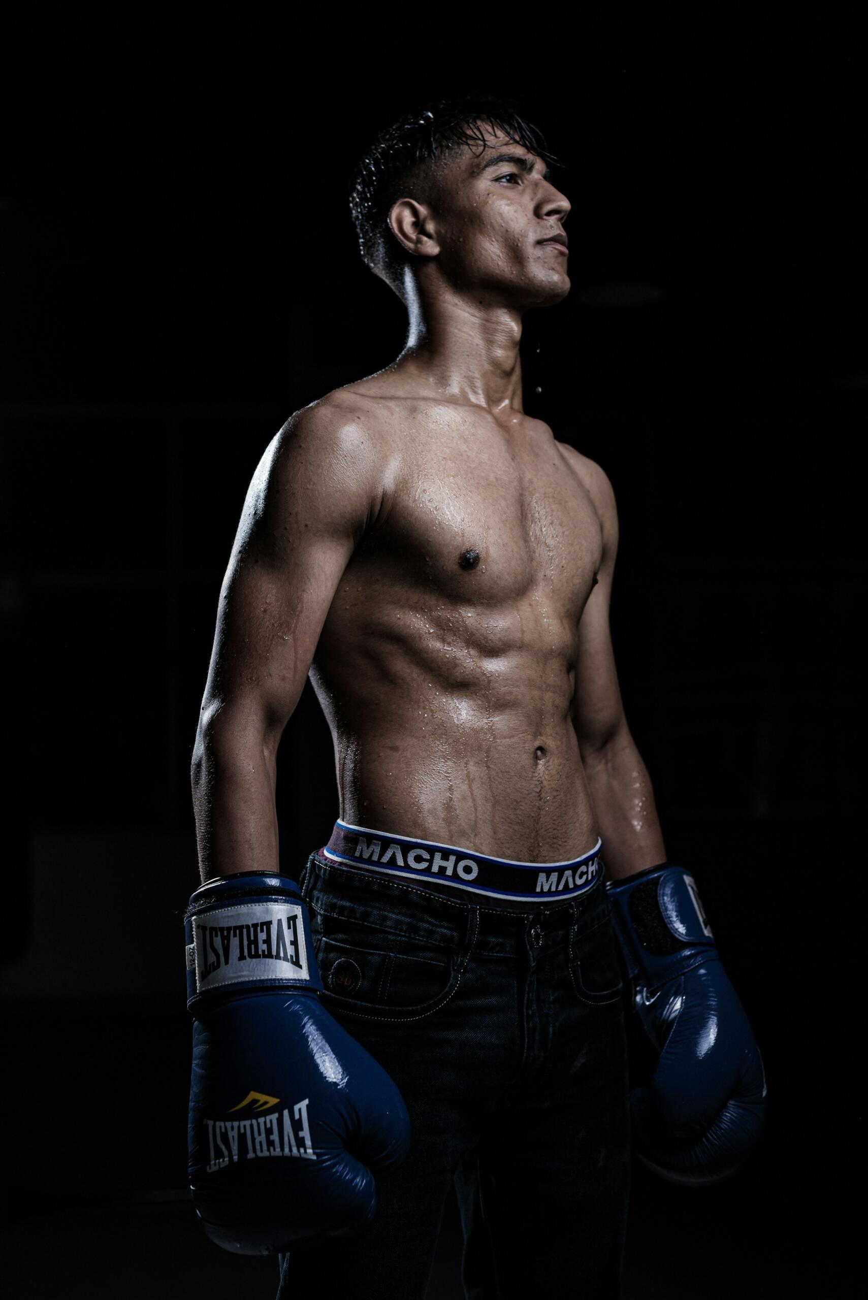 Shirtless male boxer with blue Everlast gloves and "Macho" waistband, standing in a dark, sweat-covered setting.