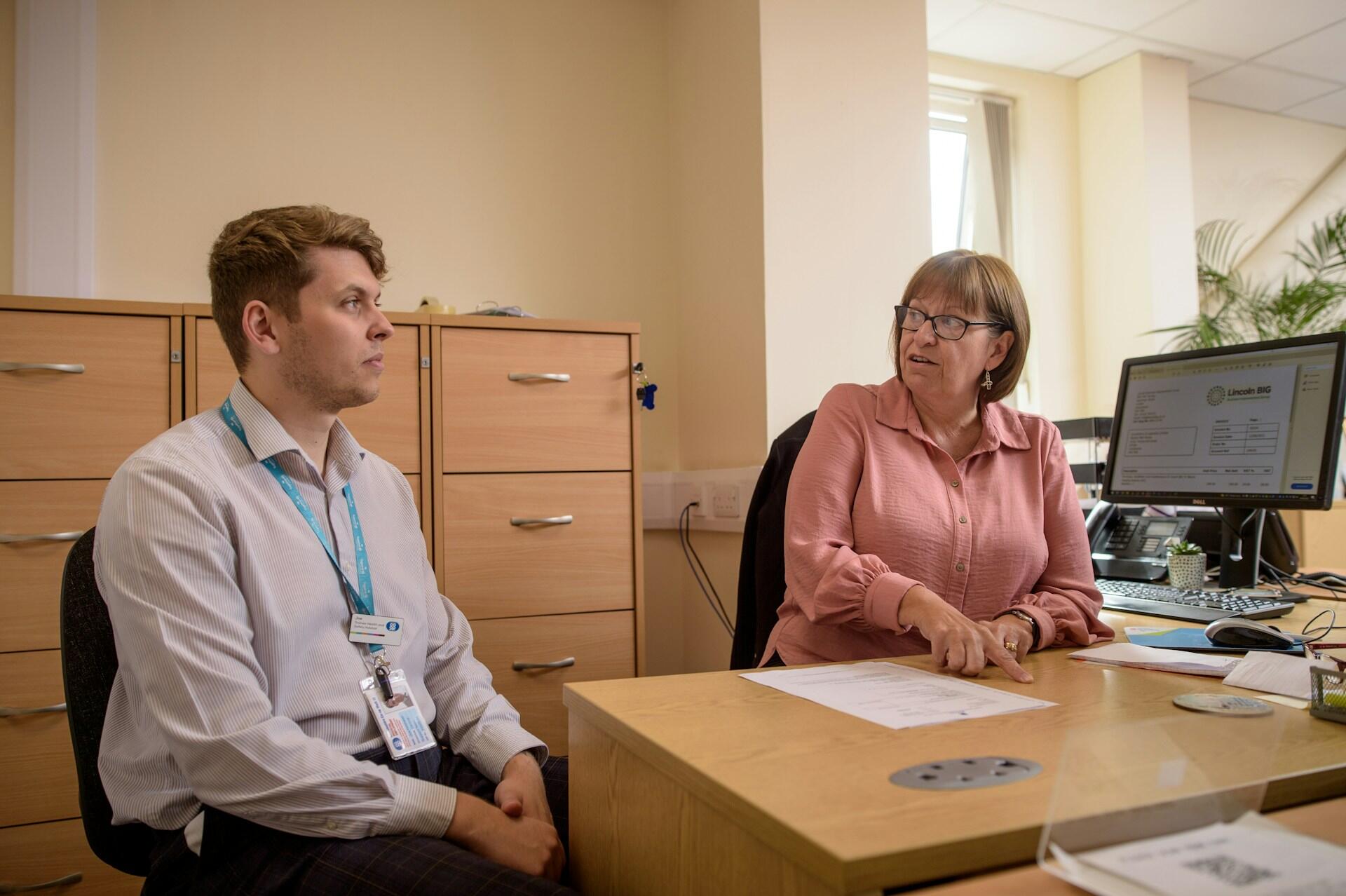 Two colleagues discussing documents in an office with a computer, filing cabinets, and natural light from a window.