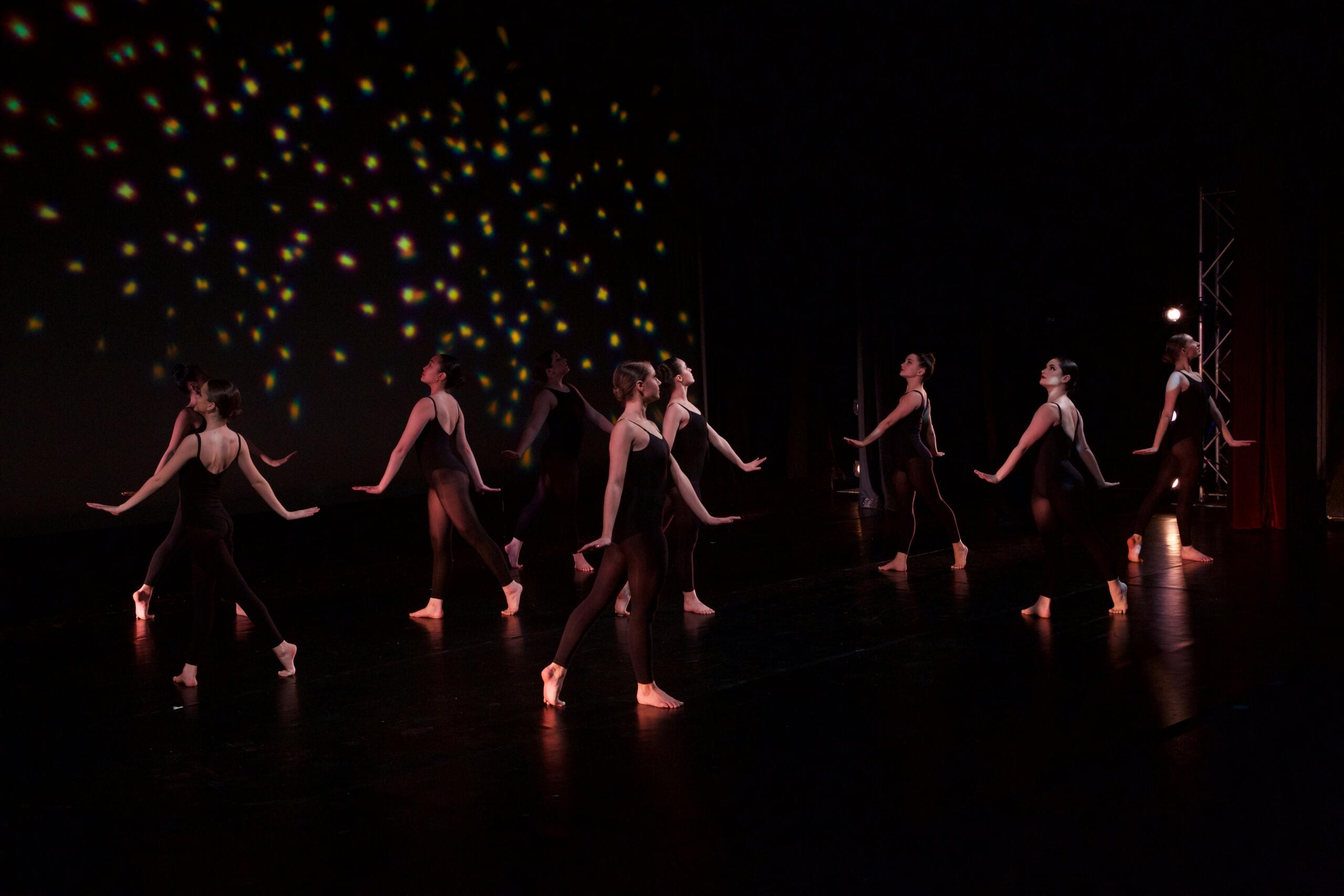 Ballet dancers in black leotards perform a synchronized routine on stage with star-like light projections in a dark theater.
