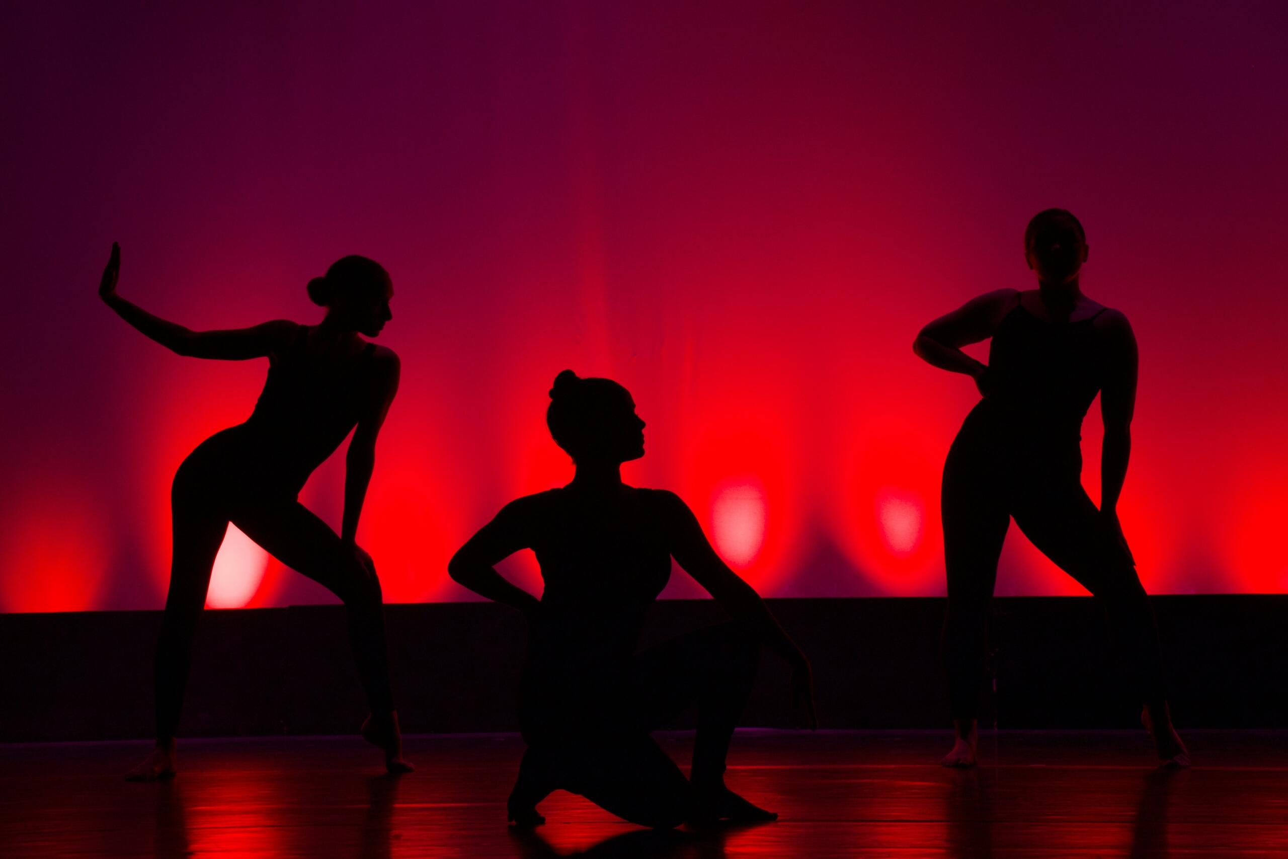 Silhouettes of three dancers posing gracefully on stage against a vibrant red and purple background light.