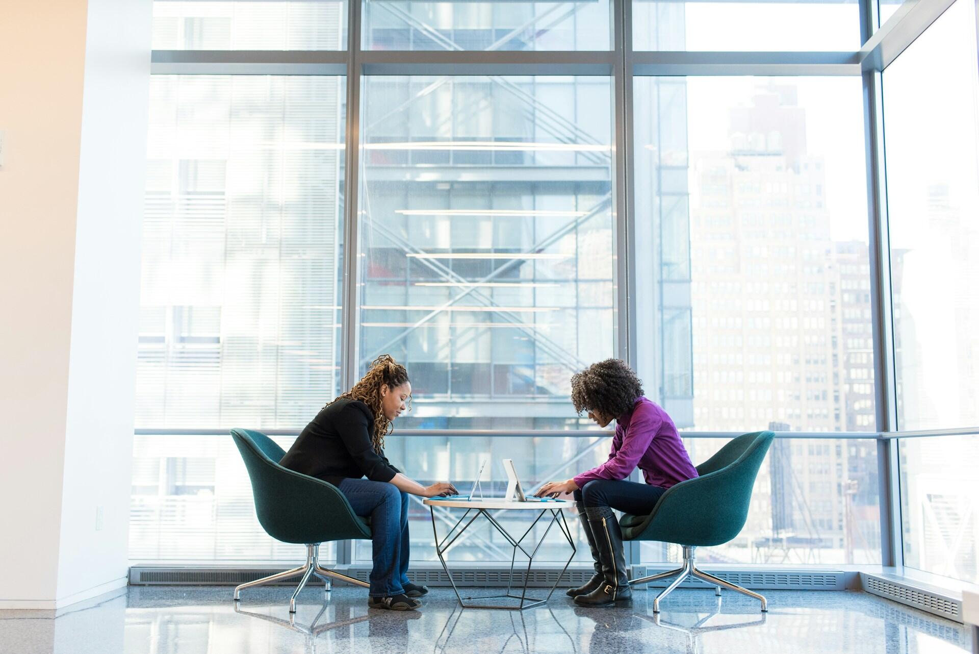 Two women seated in office chairs, engaged in conversation in a professional office setting.