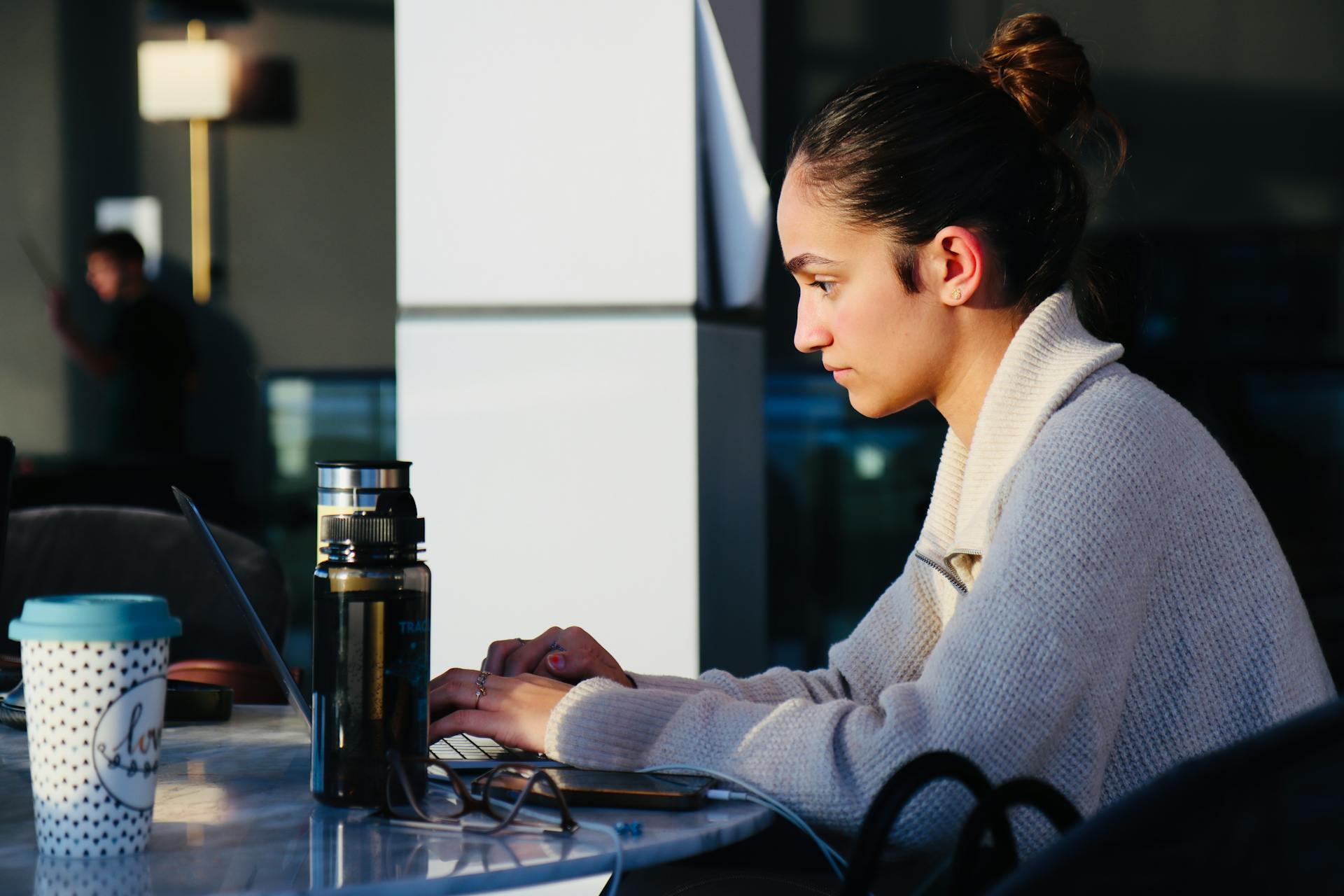 A woman sitting at a table, focused on her laptop, with a notebook and pen beside her.