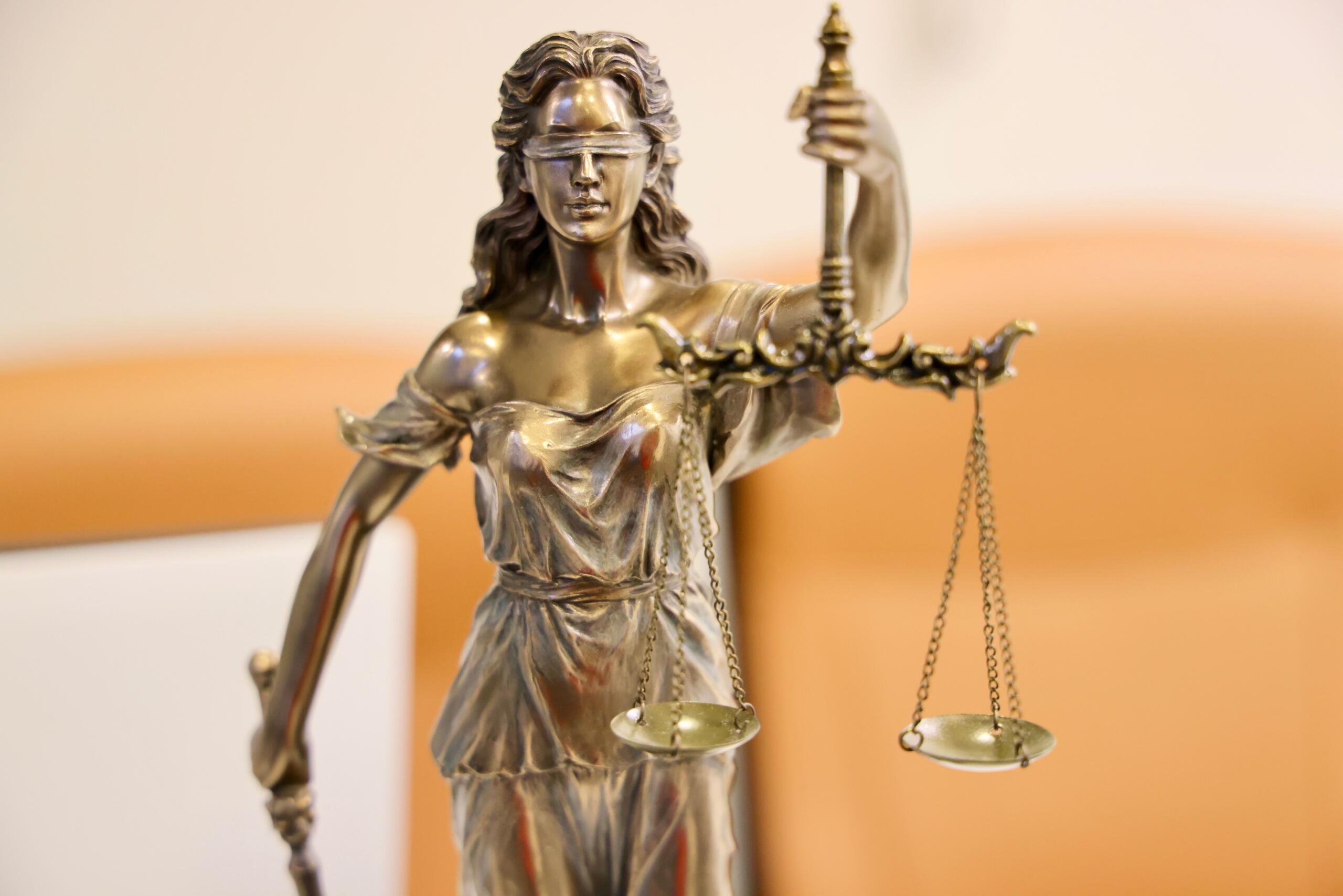 A bronze statue of Lady Justice stands holding balanced scales in one hand and a sword in the other, symbolizing fairness, impartiality, and the rule of law. She wears flowing robes, and her blindfold represents justice administered without bias or prejudice.