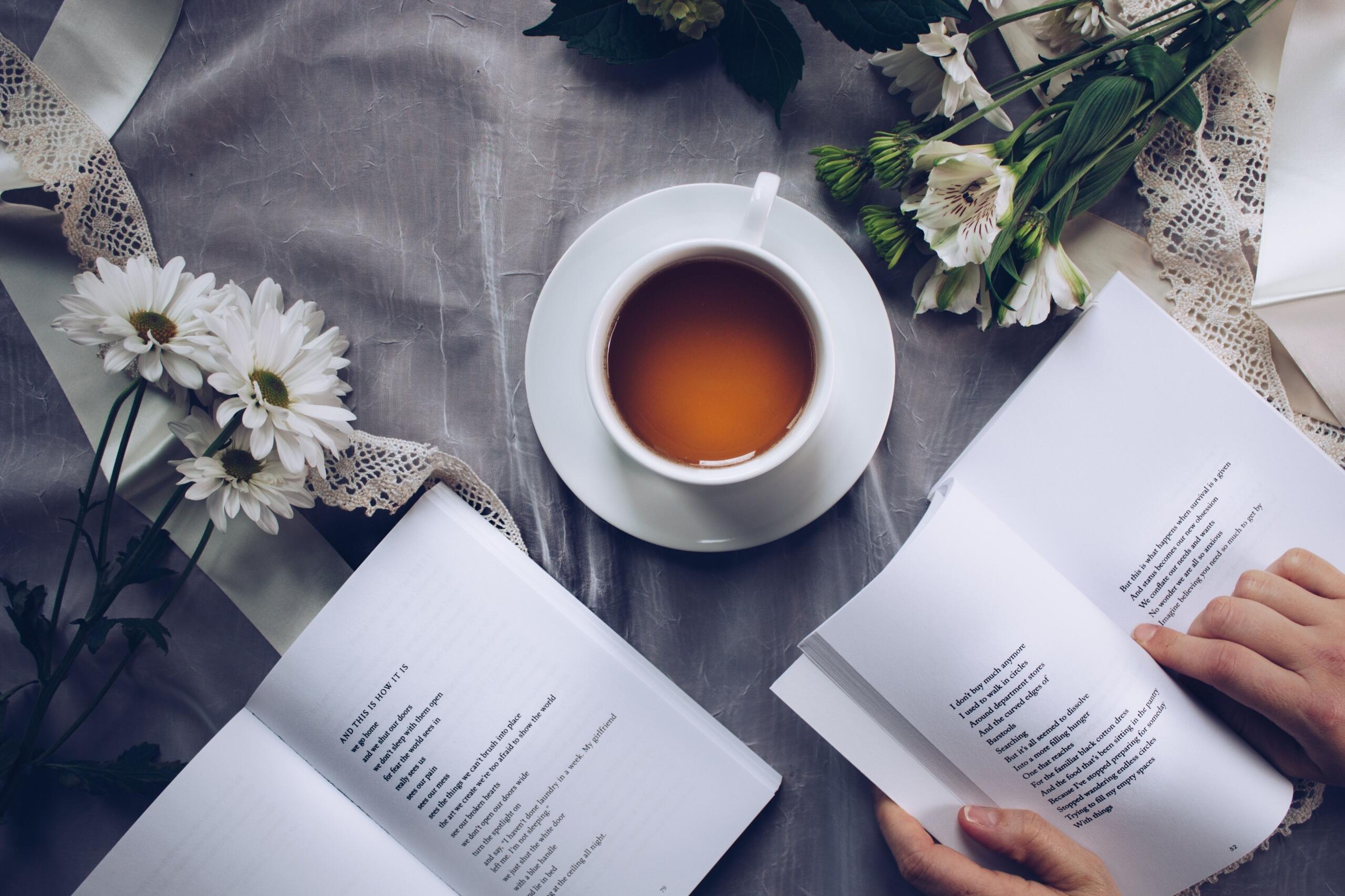 A steaming cup of tea rests on a wooden table beside several open poetry books. A person's hands are visible, holding a book and creating a quiet, cozy reading scene.