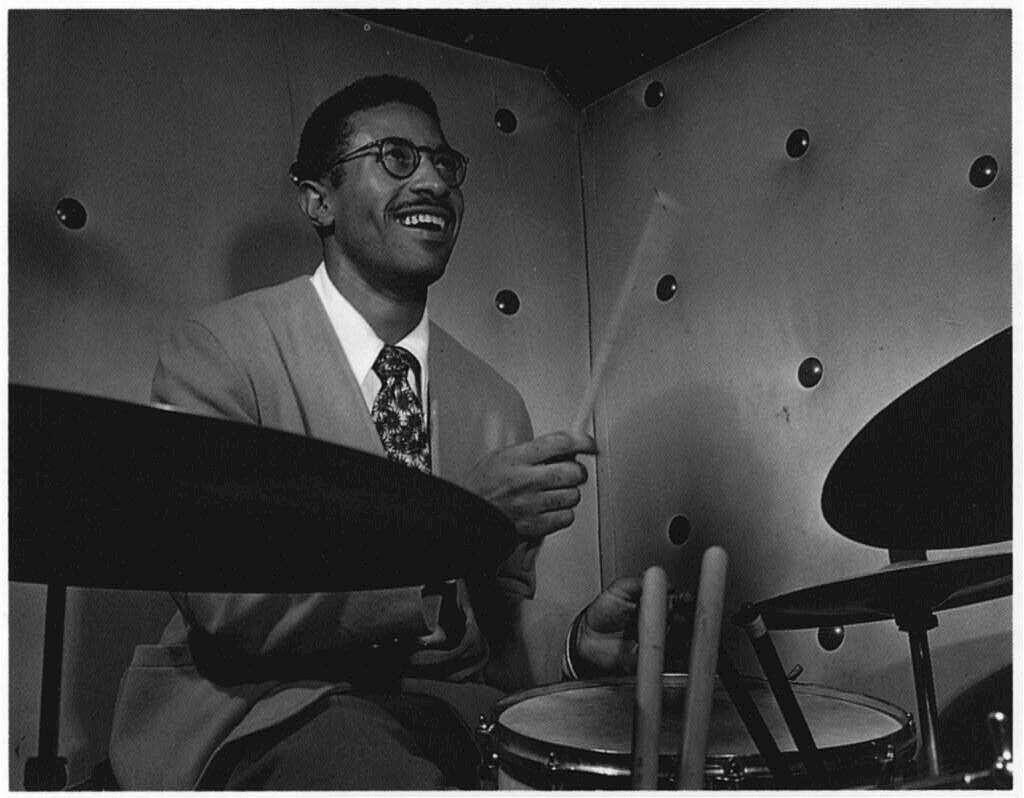 Black-and-white portrait of legendary jazz drummer Max Roach holding drumsticks while performing in a recording studio.