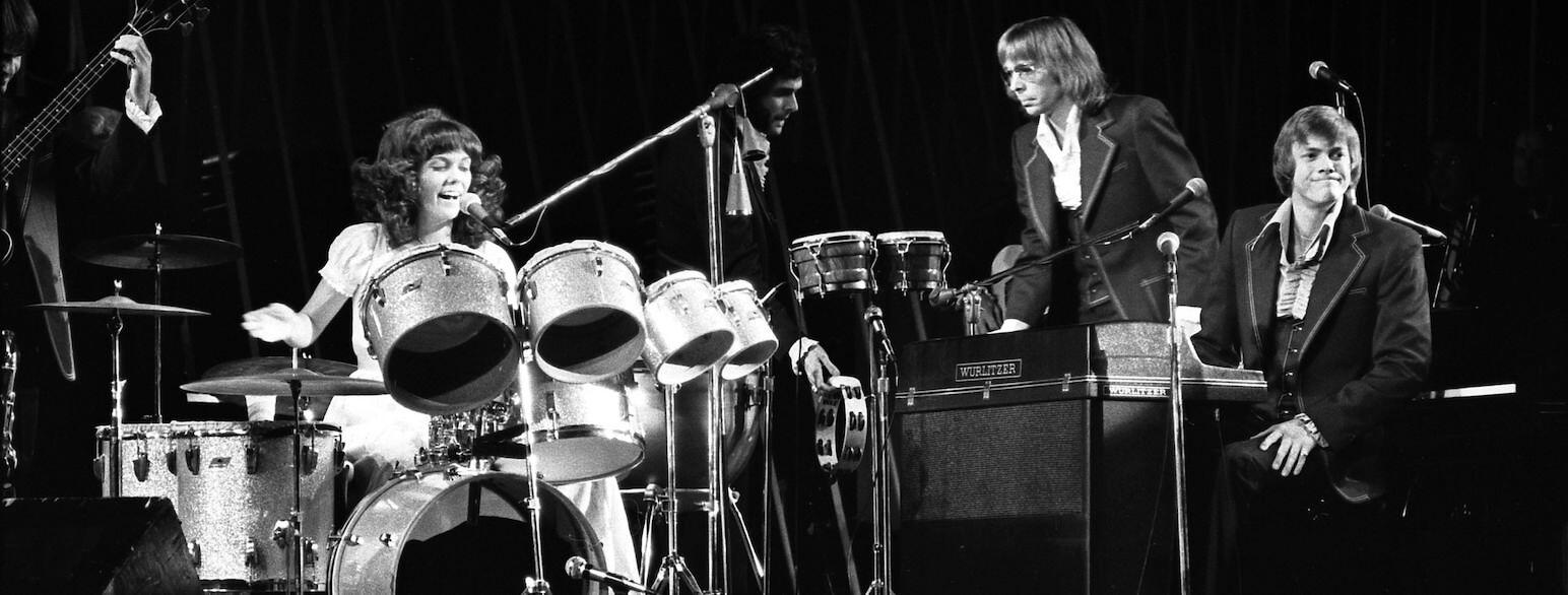 Black-and-white photo of Karen Carpenter singing and playing the drum kit with The Carpenters during a live performance at the Greek Theatre in 1975.