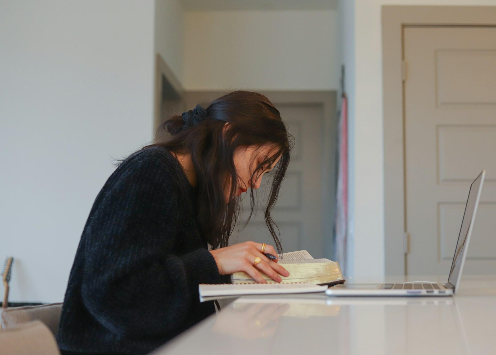 Woman studying with a book and notebook at a table, with an open laptop nearby, in a quiet indoor setting.