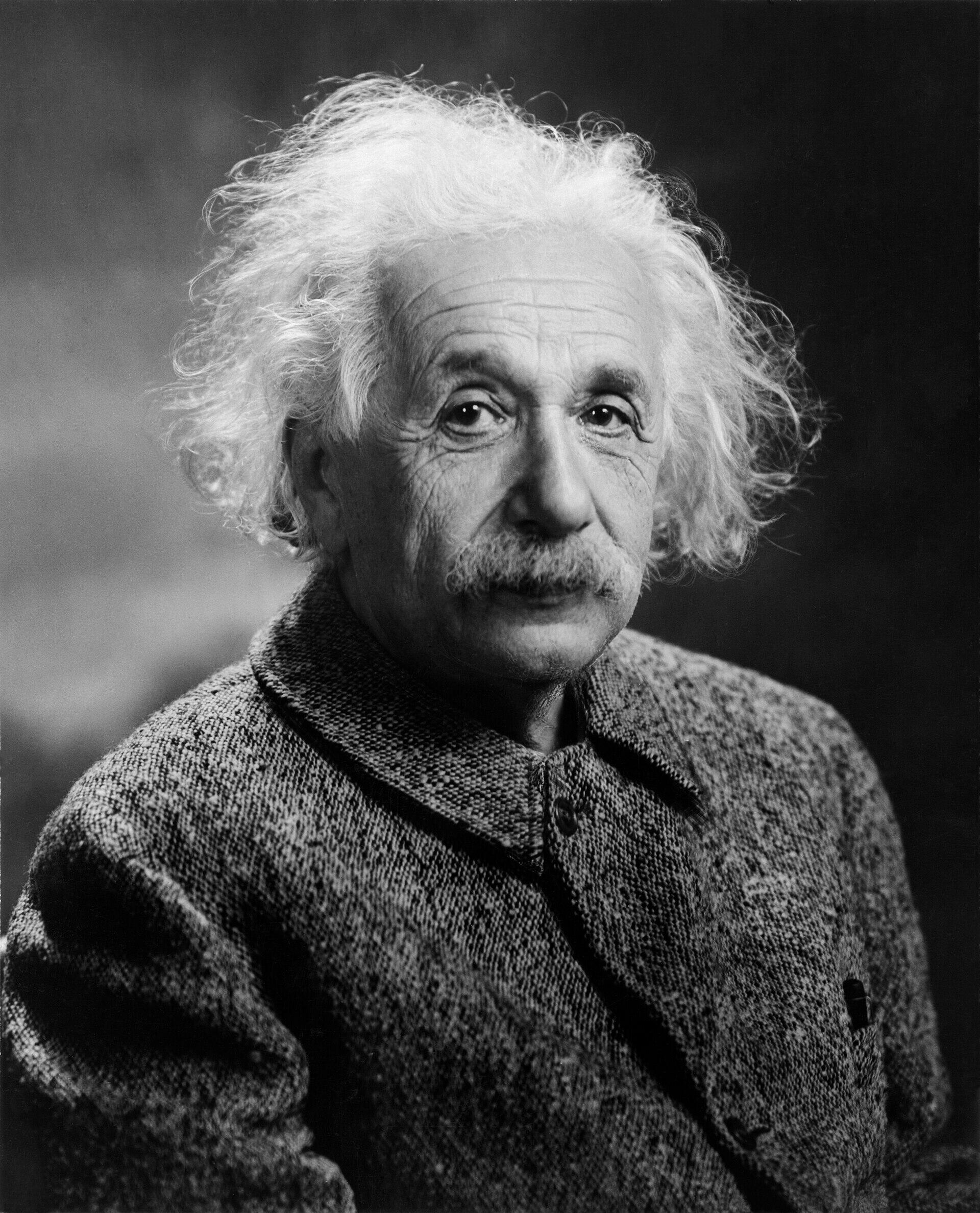 Black and white portrait of an elderly man with disheveled white hair and mustache, wearing a textured coat, against a plain dark background.