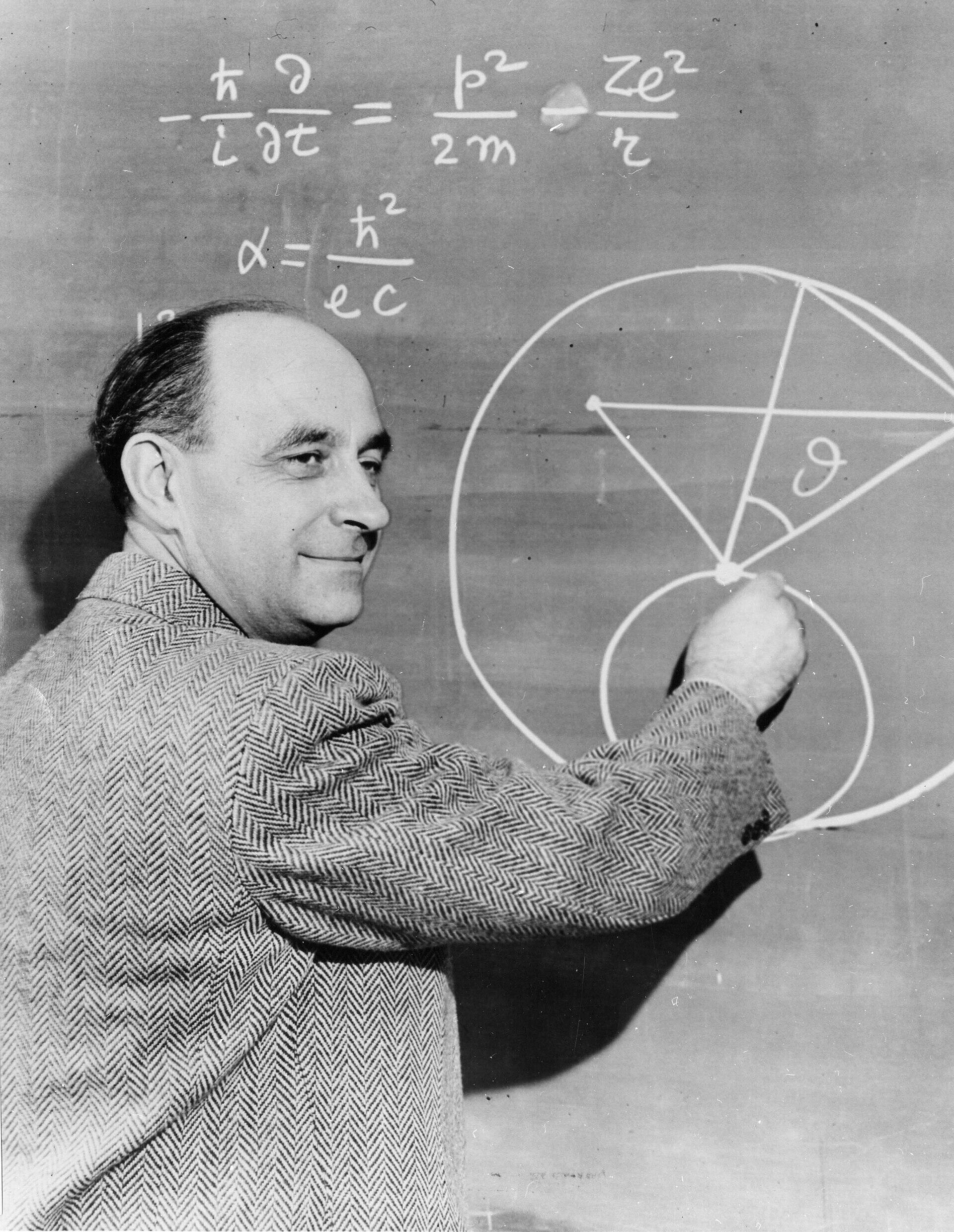 Black and white photo of a man drawing geometric and physics formulas on a chalkboard in a classroom or lecture setting.