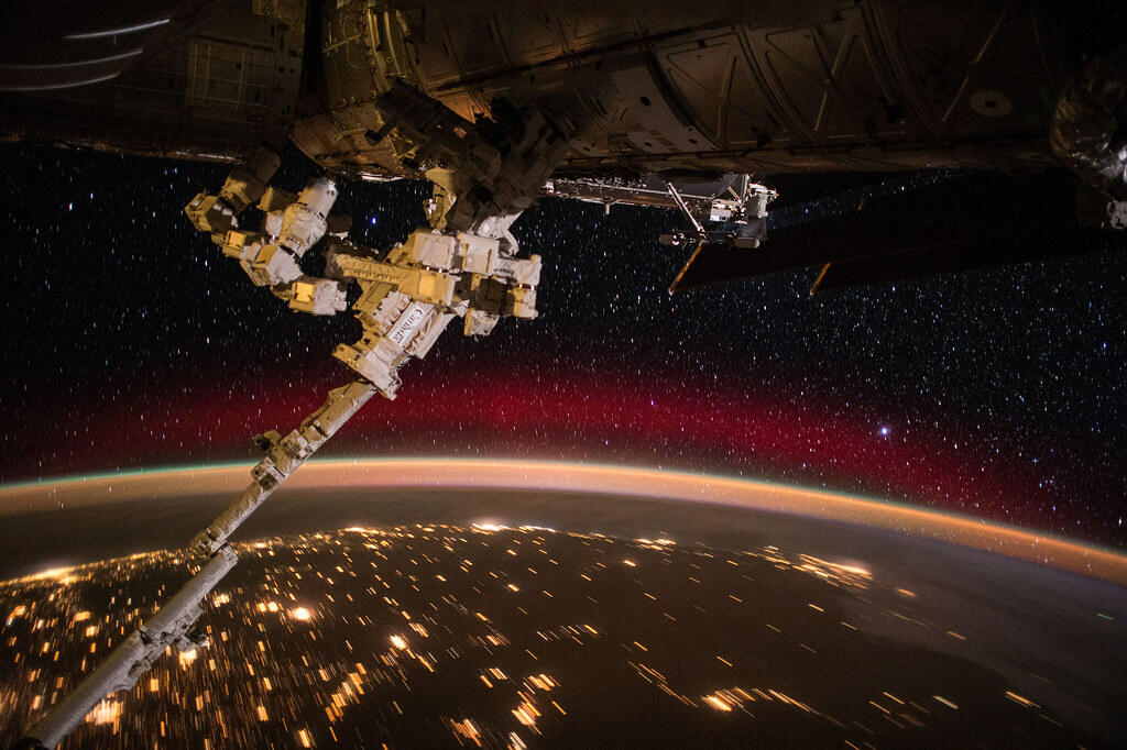 Canadarm extends from the International Space Station above Earth at night, with city lights, the planet's glowing atmosphere, and a star-filled sky visible in the background.