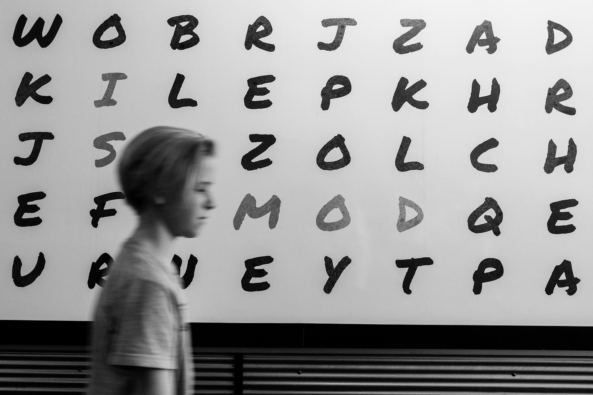 Blurred side profile of a young person walking past a wall with black and gray handwritten letters in columns.