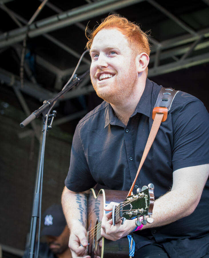 A man with short red hair plays an acoustic guitar on stage, smiling joyfully. He's wearing a black shirt, and there's a black microphone stand nearby.