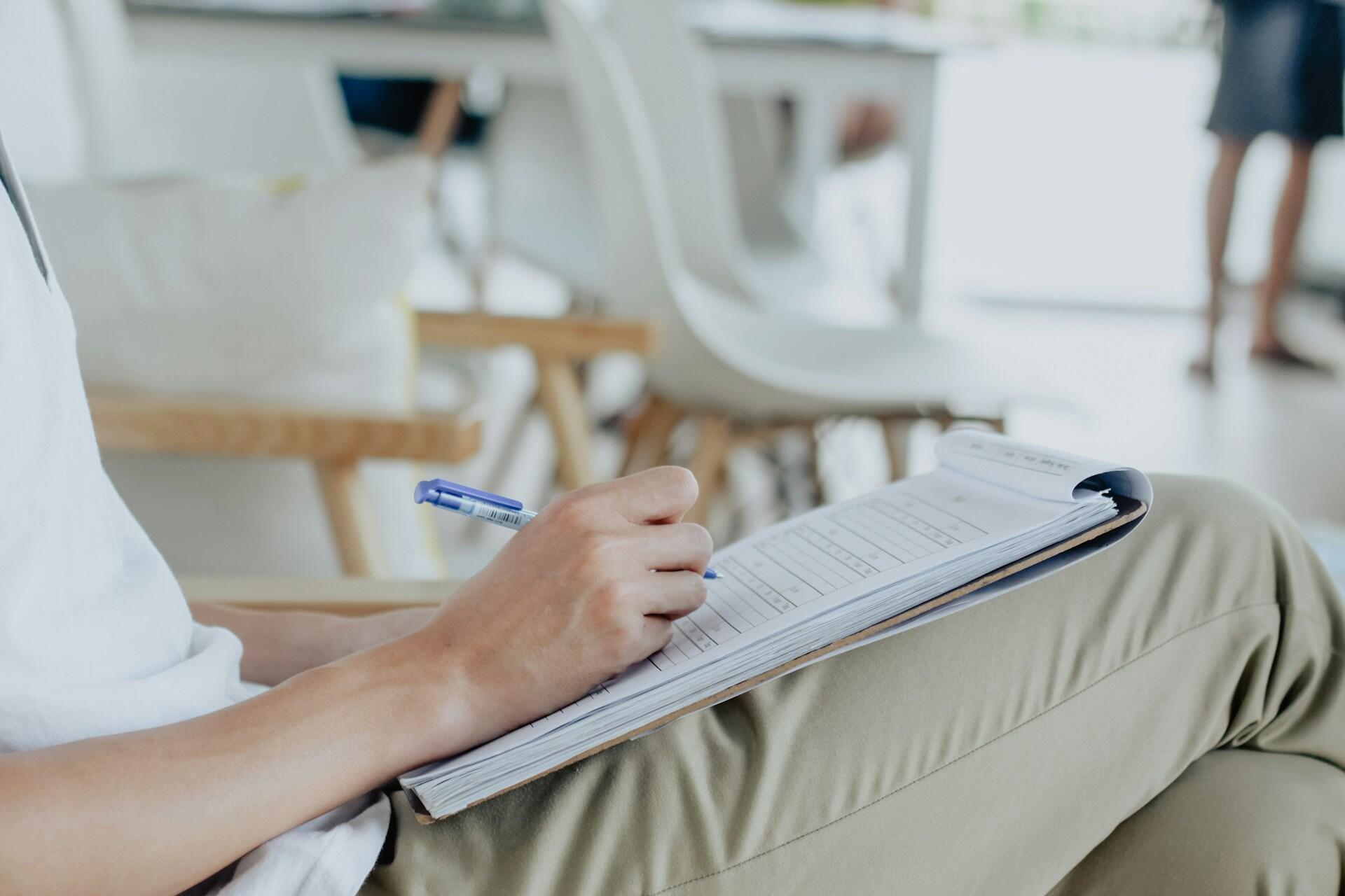 A close-up of a person's hand writing on a notebook while sitting. The person has a pen in hand and is wearing a white shirt,