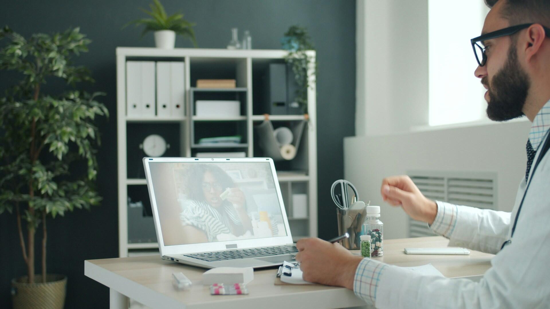 A man wearing glasses and a white shirt sits at a desk with a laptop, engaged in a video call.