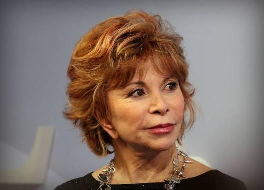 Portrait of Chilean author Isabel Allende, known for novels such as The House of the Spirits. She is shown with short auburn hair, wearing a black top and a silver necklace while speaking at a literary event.