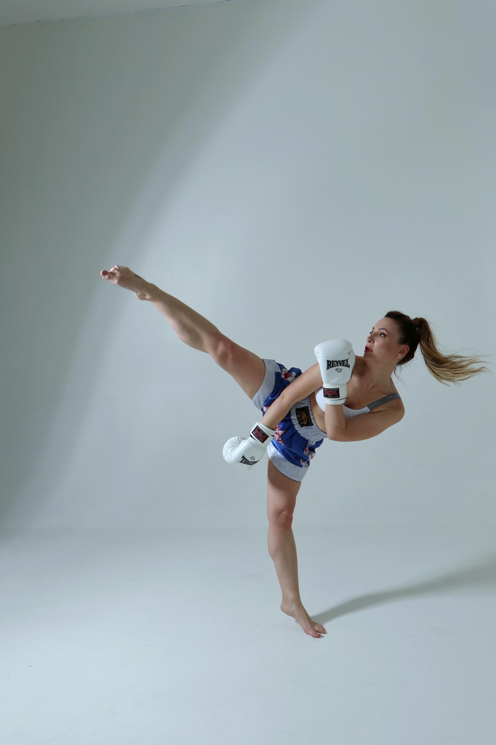 A woman wearing athletic clothing and boxing gloves practices kickboxing in a bright, all-white training room. Source: Skoro Photo.