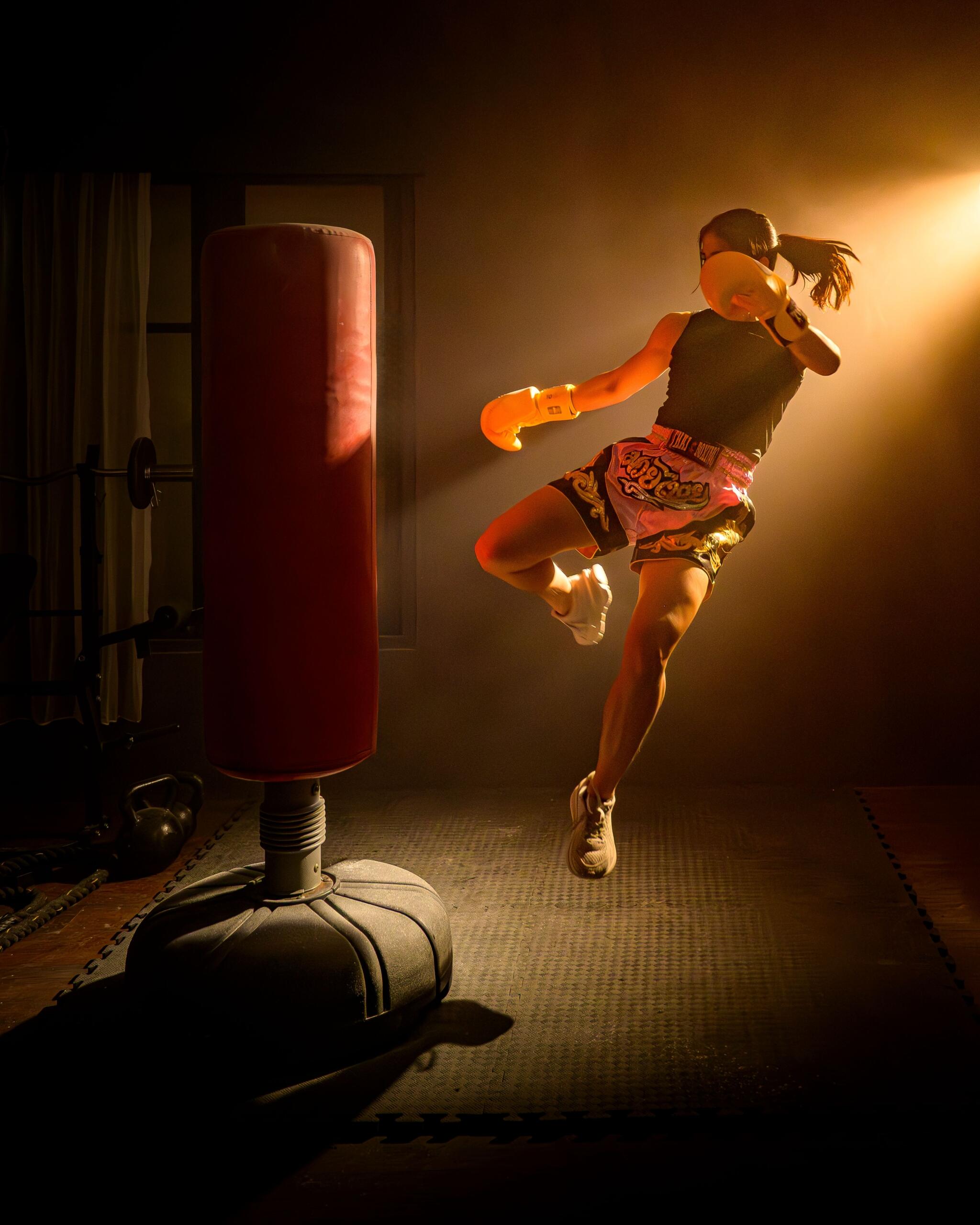A woman wearing athletic clothing and boxing gloves trains with a heavy punching bag in a gym. She is captured mid-punch in a balanced fighting stance, demonstrating focus, strength, and proper kickboxing technique. The training space includes fitness equipment, emphasizing an active workout environment. Source: Pexels.