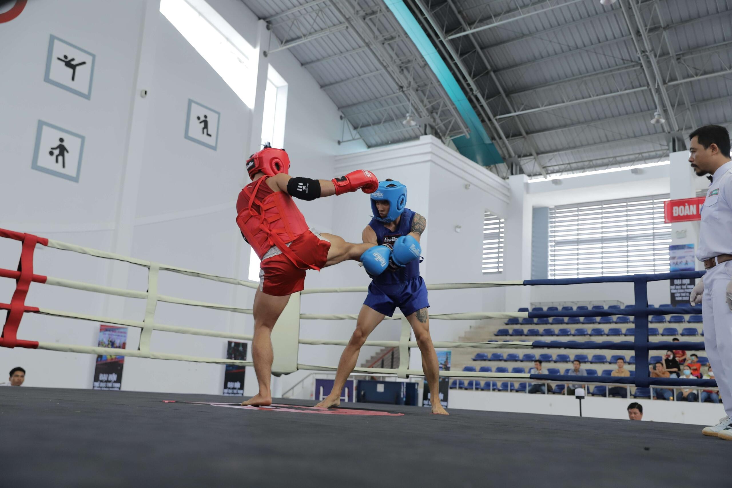 Two men compete in a Muay Thai match inside a boxing ring, wearing boxing gloves, shorts, and protective gear. One fighter throws a strike while the other defends, showcasing the sport's fast-paced action and athleticism. Bright arena lights illuminate the ring as a referee and spectators watch the bout.