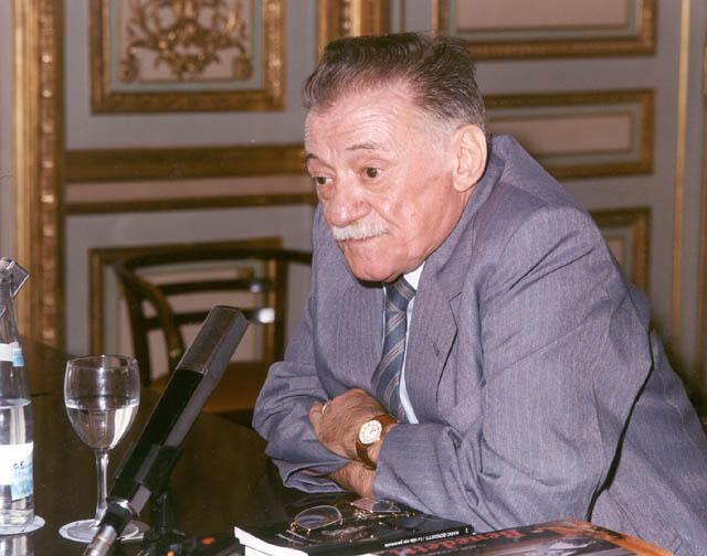 Photograph of Uruguayan writer and poet Mario Benedetti seated at a table during a literary event. Wearing a gray suit, he leans toward a microphone while speaking, with books, a glass of water, and ornate interior décor in the background.