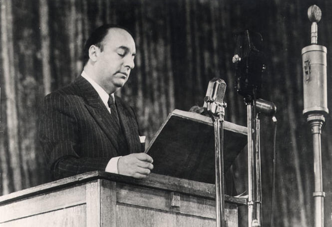 Black-and-white photograph of Chilean poet and Nobel Prize laureate Pablo Neruda delivering a speech from a podium, reading from a manuscript into several microphones during a public event.