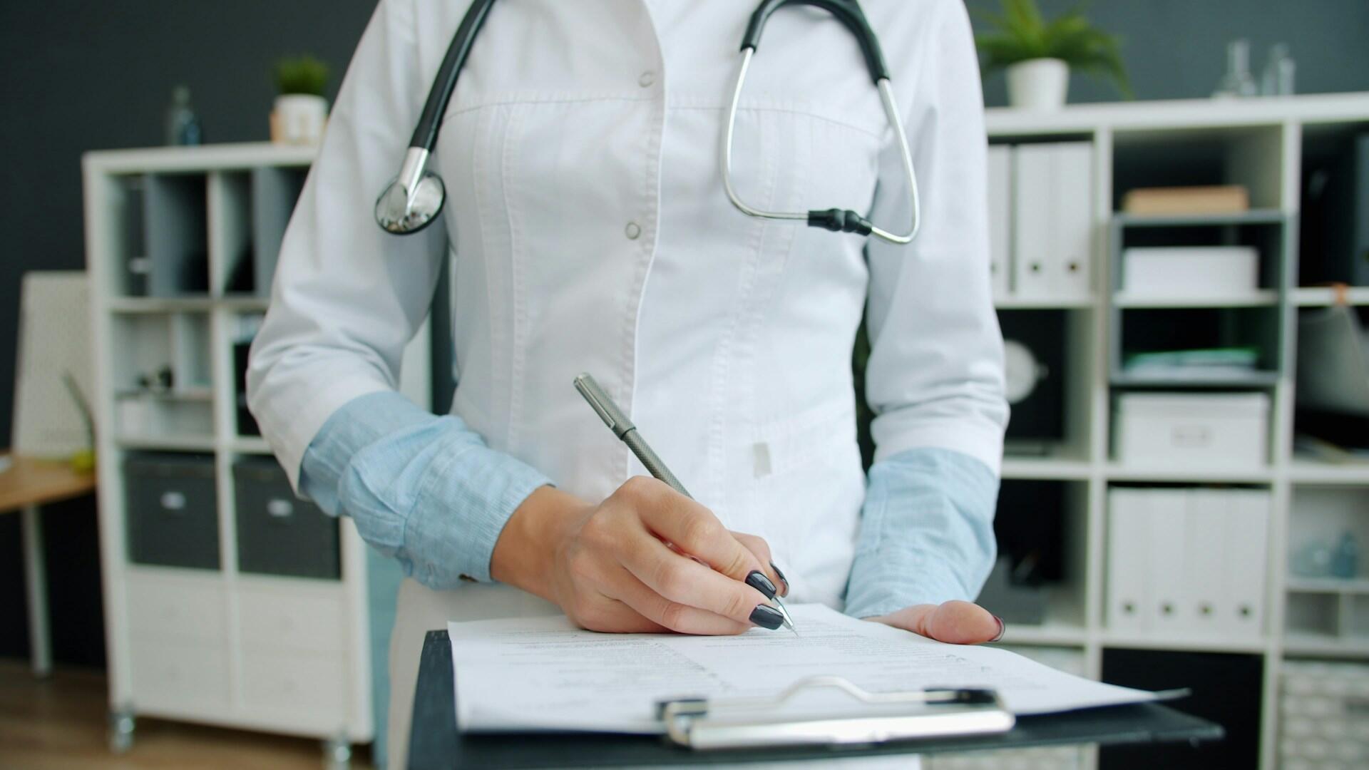 A female doctor in a white coat writes notes on a clipboard, focused on her task in a clinical setting.
