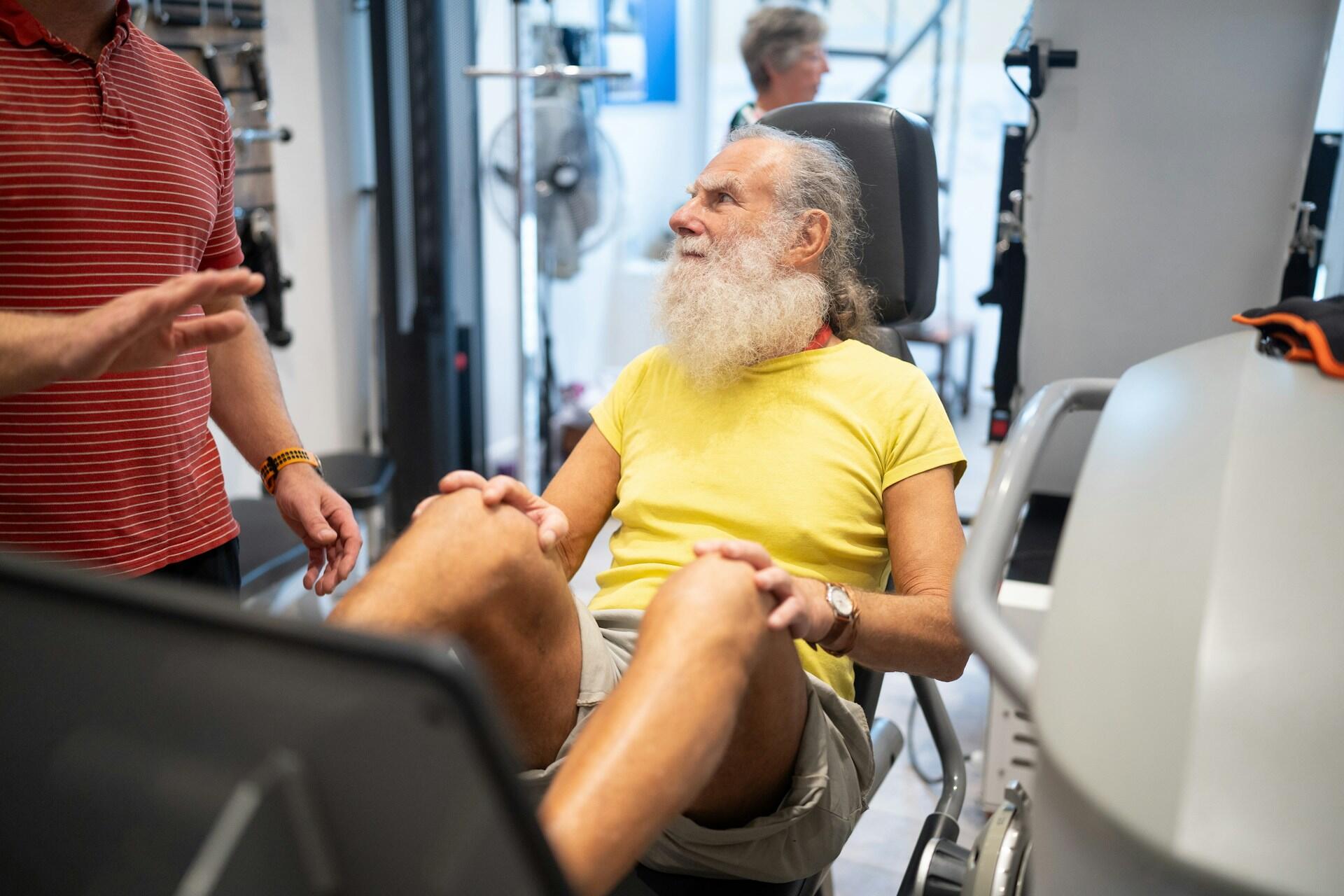 A bearded man sitting on a chair in a gym, surrounded by fitness equipment and weights.